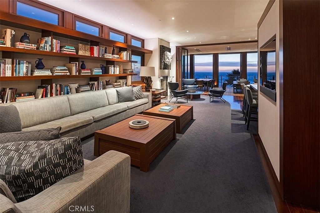 This sophisticated living room features a long, neutral-toned sofa paired with two matching wooden coffee tables, creating a balanced seating area. A wall of built-in shelving provides ample storage and display space, while large windows offer a stunning, unobstructed view of the ocean at sunset. The room is finished with dark carpeting and warm wood accents, leading toward an open-concept dining area and a deck beyond.