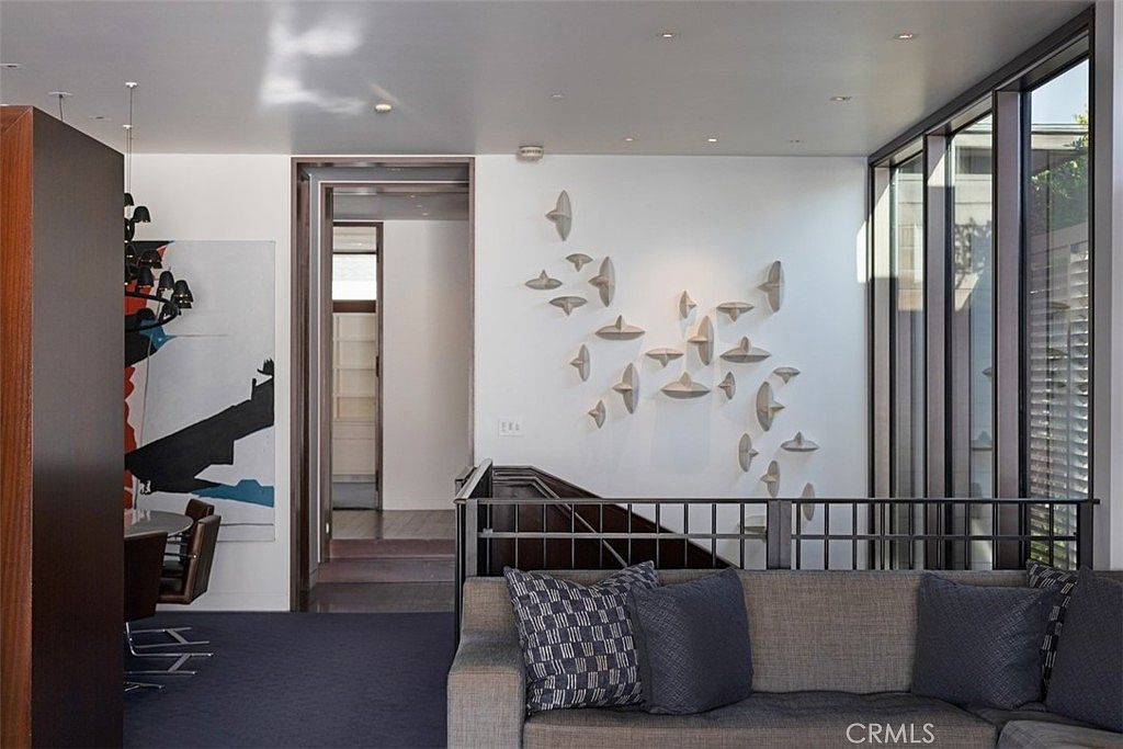 This contemporary living space features a neutral-toned sofa adorned with patterned throw pillows, positioned against a wall decorated with a unique, sculptural art installation. To the right, large windows provide ample natural light, while a dark-toned railing separates the seating area from a staircase. A doorway leads to a secondary room, and a glimpse of a dining area with a modern chandelier is visible on the left, creating an open and sophisticated atmosphere.