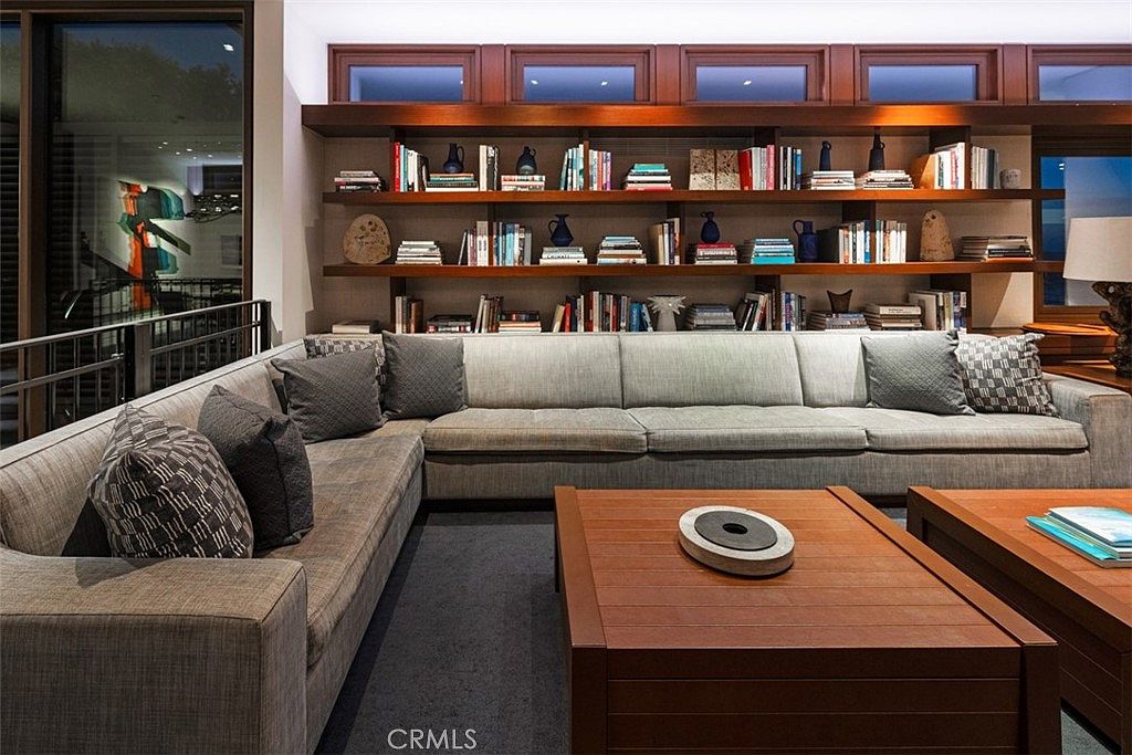 This sophisticated living room features a large, L-shaped sectional sofa upholstered in a neutral fabric, centered around a substantial wooden coffee table. A wall-to-wall built-in shelving unit displays a curated collection of books and decorative objects, illuminated by integrated lighting. The space is framed by large windows on the left, offering a view of the exterior, and maintains a warm, contemporary aesthetic.