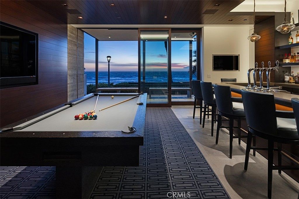 This sophisticated game room features a professional-grade pool table as the centerpiece, set against a backdrop of dark wood-paneled walls and a bar area with high-top seating. Large glass sliding doors provide an expansive, unobstructed view of the ocean at twilight, seamlessly blending indoor entertainment with the coastal landscape. The space is defined by a geometric patterned rug, recessed ceiling lighting, and a sleek, modern aesthetic perfect for evening relaxation.