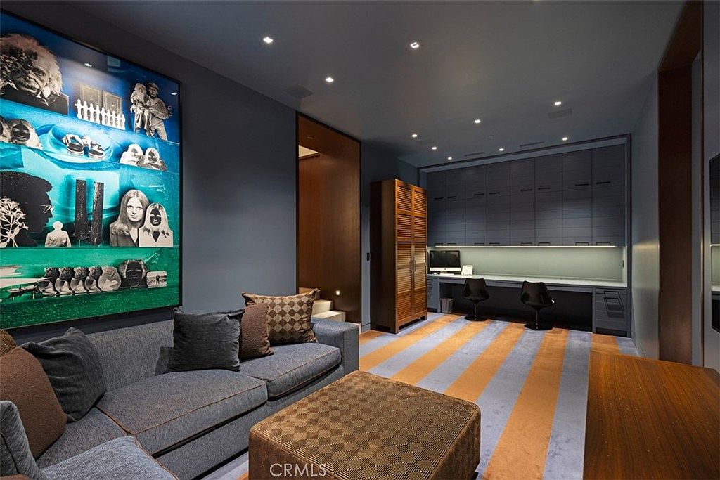 This contemporary office and lounge space features a large grey sofa with patterned accent pillows and a matching ottoman, positioned across from a built-in desk area. The room is defined by a bold, striped carpet in blue and orange, complemented by dark blue walls and a large, vibrant art piece. A tall wooden storage cabinet stands near the desk, which is equipped with two chairs and integrated cabinetry above.