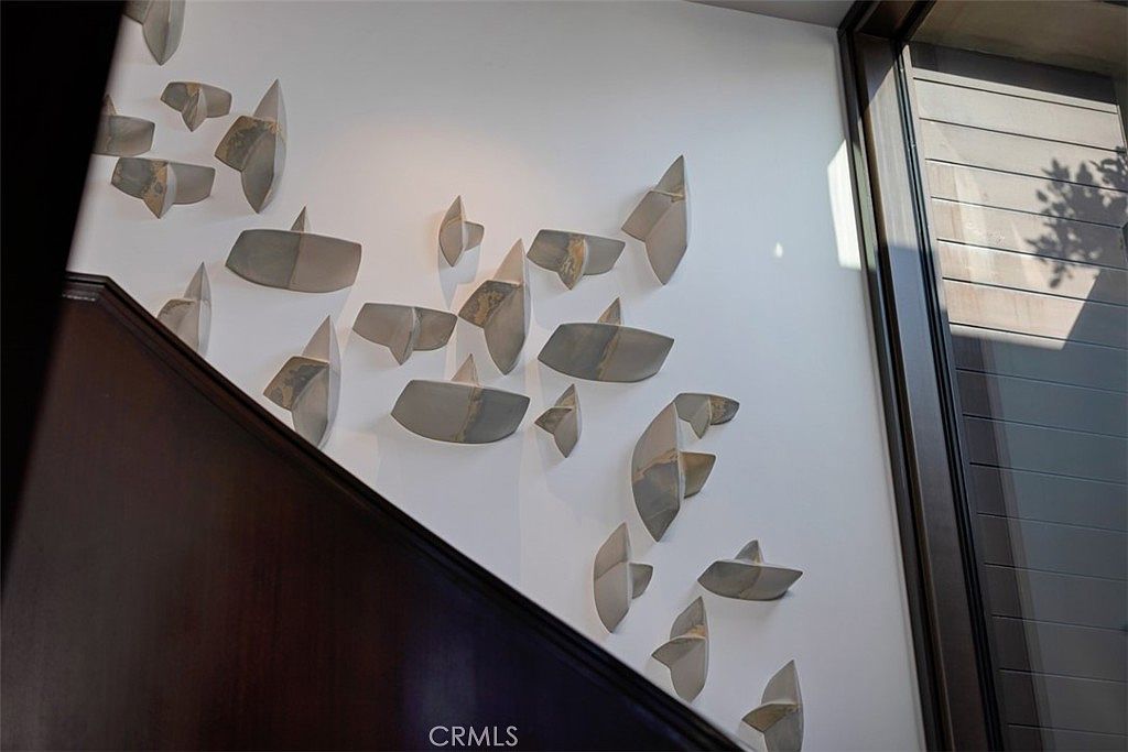 This image captures a striking wall installation featuring numerous abstract, metallic sculptural elements arranged in a dynamic, floating pattern against a crisp white wall. The composition is framed by a dark, angled wooden staircase railing on the left and a tall window structure on the right, creating a sophisticated and artistic focal point. The perspective is angled upward, emphasizing the height and modern aesthetic of the architectural space.