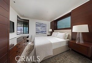 This modern primary bedroom features a clean, minimalist aesthetic with dark wood paneling on the walls and a neutral-toned bed. A large window above the headboard allows natural light to filter in, while a sleek dresser and a bedside lamp with a white shade complete the space. The room is grounded by a textured area rug, creating a sophisticated and tranquil atmosphere.