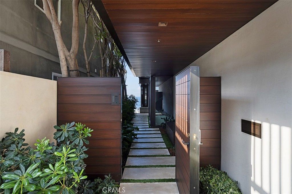 This modern entryway features a wide, horizontal-slat wood gate that opens to a stone-paved walkway lined with lush greenery. A deep, dark wood-paneled ceiling extends over the path, creating a dramatic, tunnel-like perspective that draws the eye toward the ocean view in the distance. The clean lines and natural materials provide a sophisticated, contemporary aesthetic for this private home entrance.