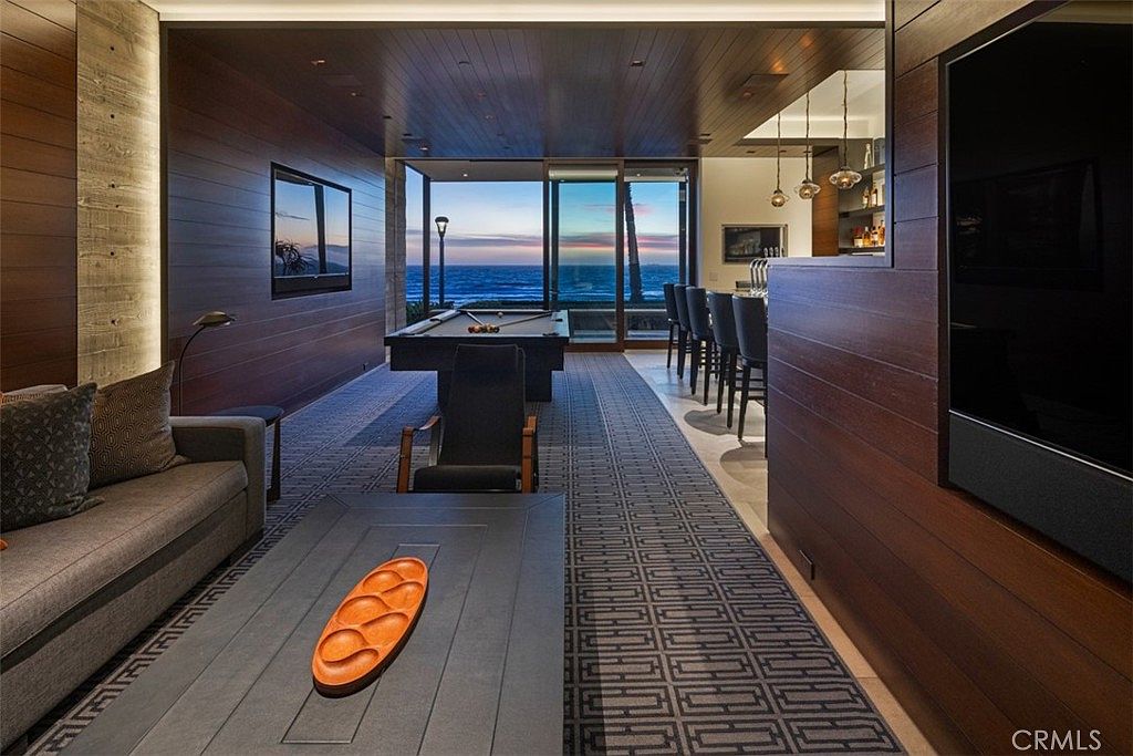 This sophisticated interior space functions as a multi-purpose lounge and game room, featuring dark wood-paneled walls and a patterned area rug. The room includes a pool table positioned in front of large glass sliding doors that offer a panoramic view of the ocean at sunset, alongside a comfortable seating area with a sofa and a central coffee table. To the right, a bar area with high stools and pendant lighting completes the entertainment-focused layout.