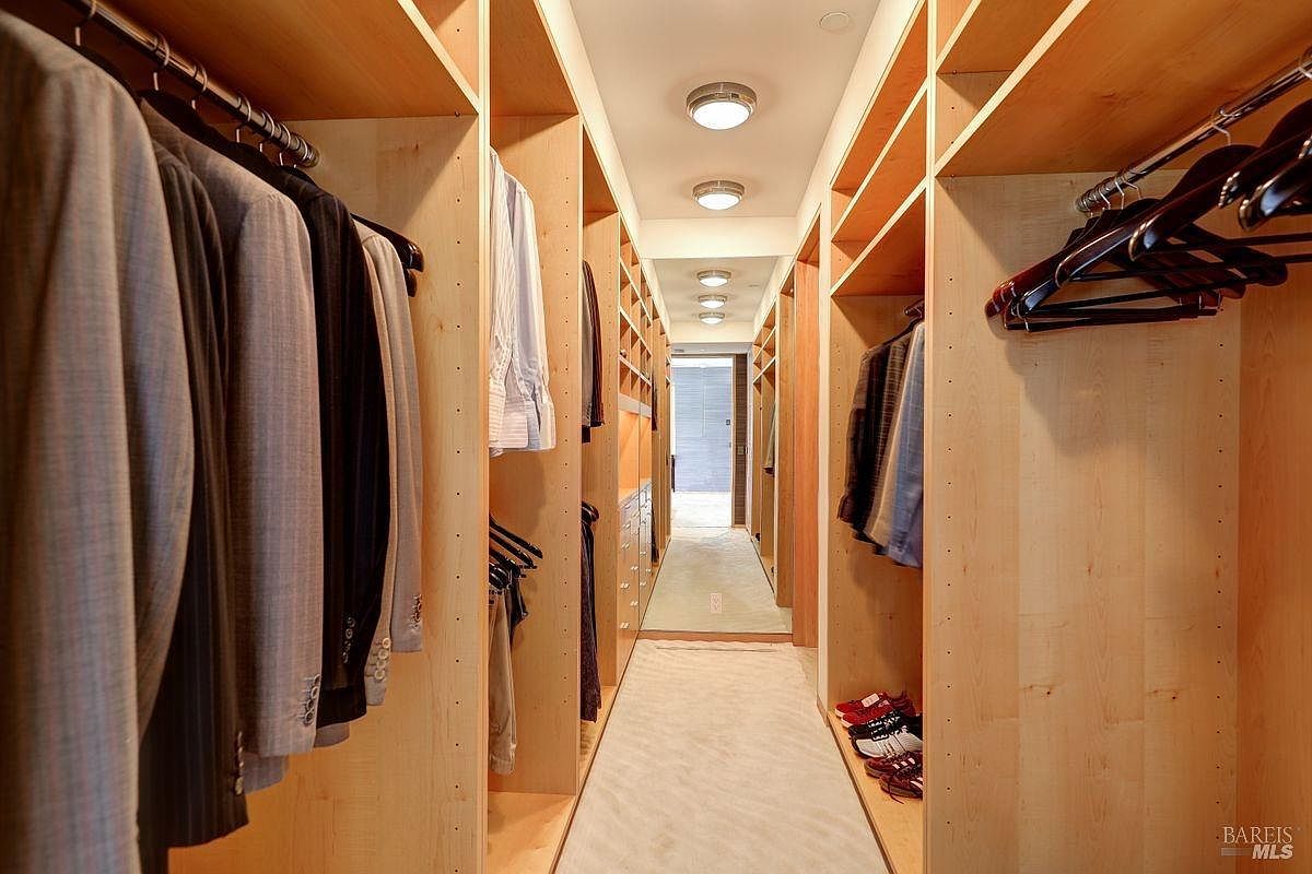 This spacious walk-in closet features extensive light wood cabinetry with integrated hanging rods and shelving on both sides. The narrow, elongated layout is illuminated by a series of recessed ceiling lights, leading the eye toward a bright doorway at the far end. A neutral-toned carpet runs throughout the space, creating a clean and organized aesthetic suitable for a high-end primary suite.