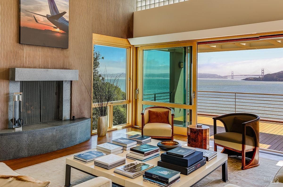 This sophisticated living room features a stone-clad fireplace and a large coffee table stacked with books, serving as the central focal point. The space is defined by its expansive glass walls that offer panoramic views of the bay and the Golden Gate Bridge, seamlessly connecting the interior to the outdoor deck. The room is furnished with elegant armchairs and warm-toned wood paneling, creating a luxurious and serene atmosphere.