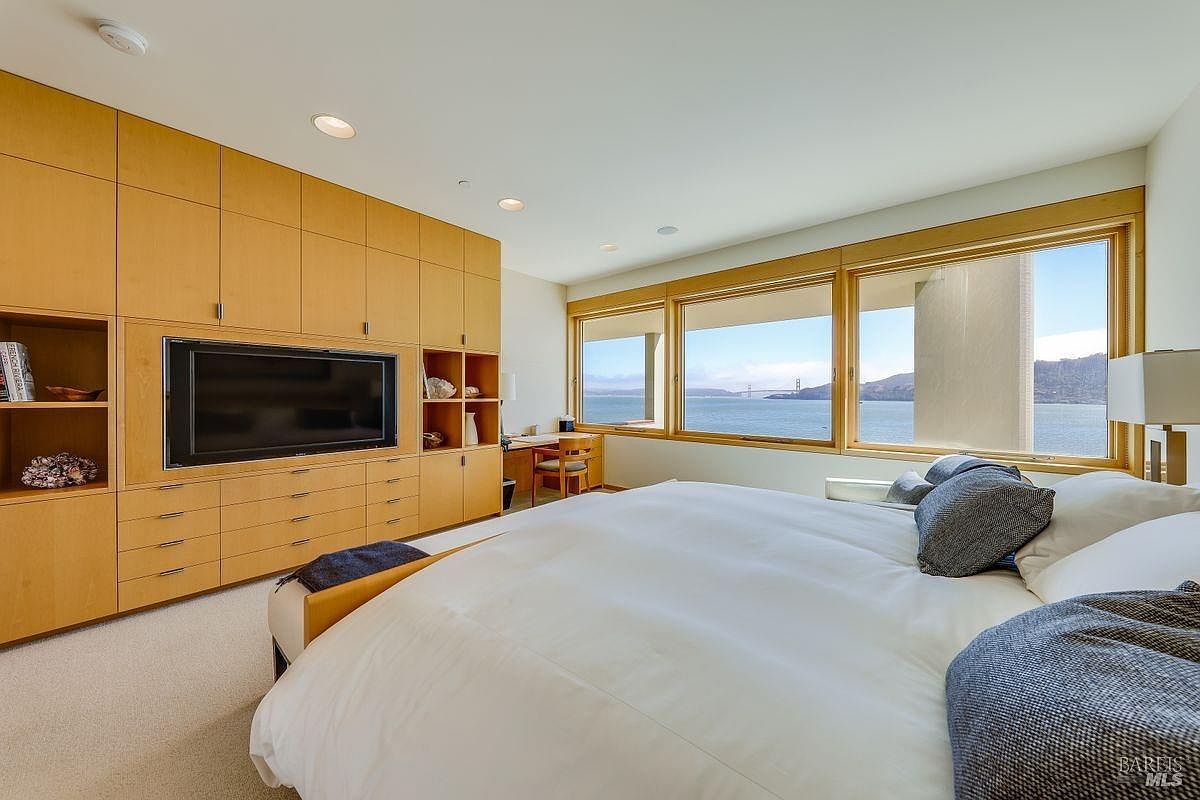 This primary bedroom features a large, light wood built-in wall unit that integrates a television and shelving, providing ample storage. A plush bed is positioned to face a wall of expansive windows that offer a panoramic view of the bay and the Golden Gate Bridge. The room is bright and airy, with a neutral color palette and a small desk area tucked into the corner near the window.