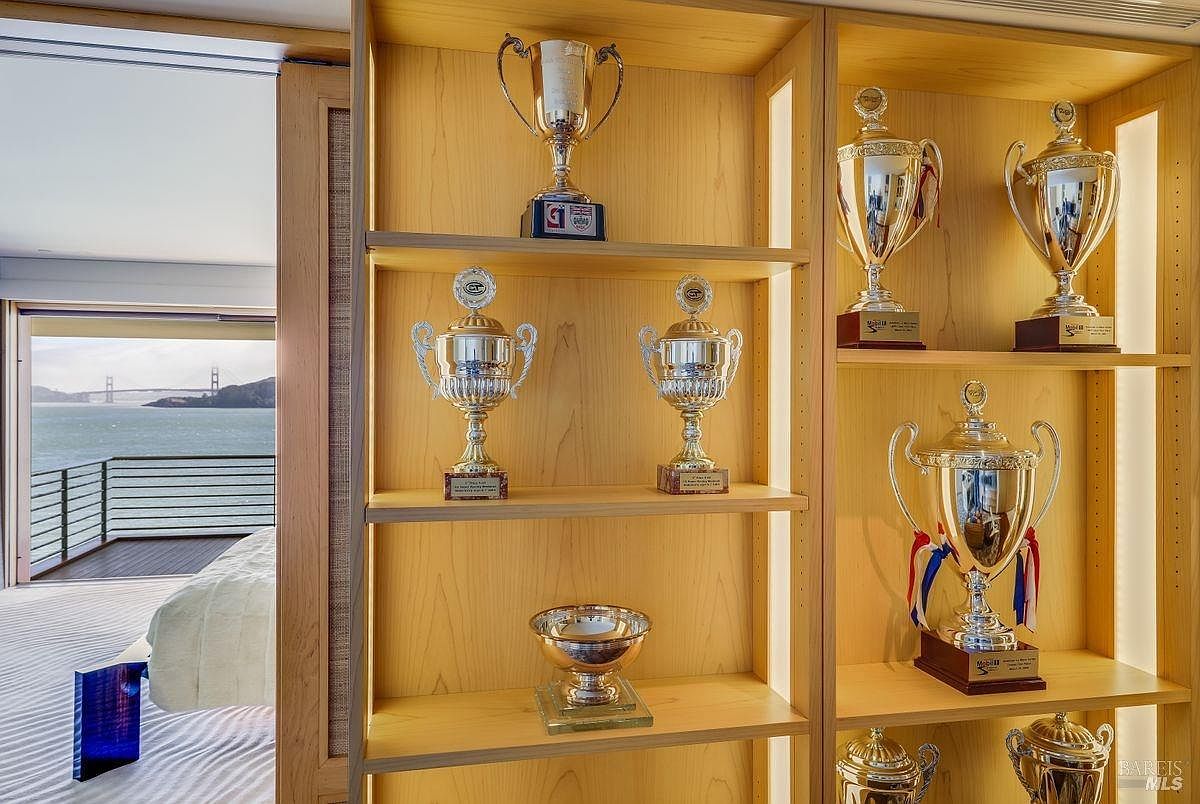 This image captures a custom-built light wood shelving unit filled with an extensive collection of silver and gold trophies, serving as a focal point within a bedroom. To the left, a sliding glass door opens onto a private balcony, revealing a stunning, unobstructed view of the Golden Gate Bridge across the water. The perspective is from inside the room, looking past the display case toward the scenic outdoor vista, creating a sophisticated and personalized atmosphere.