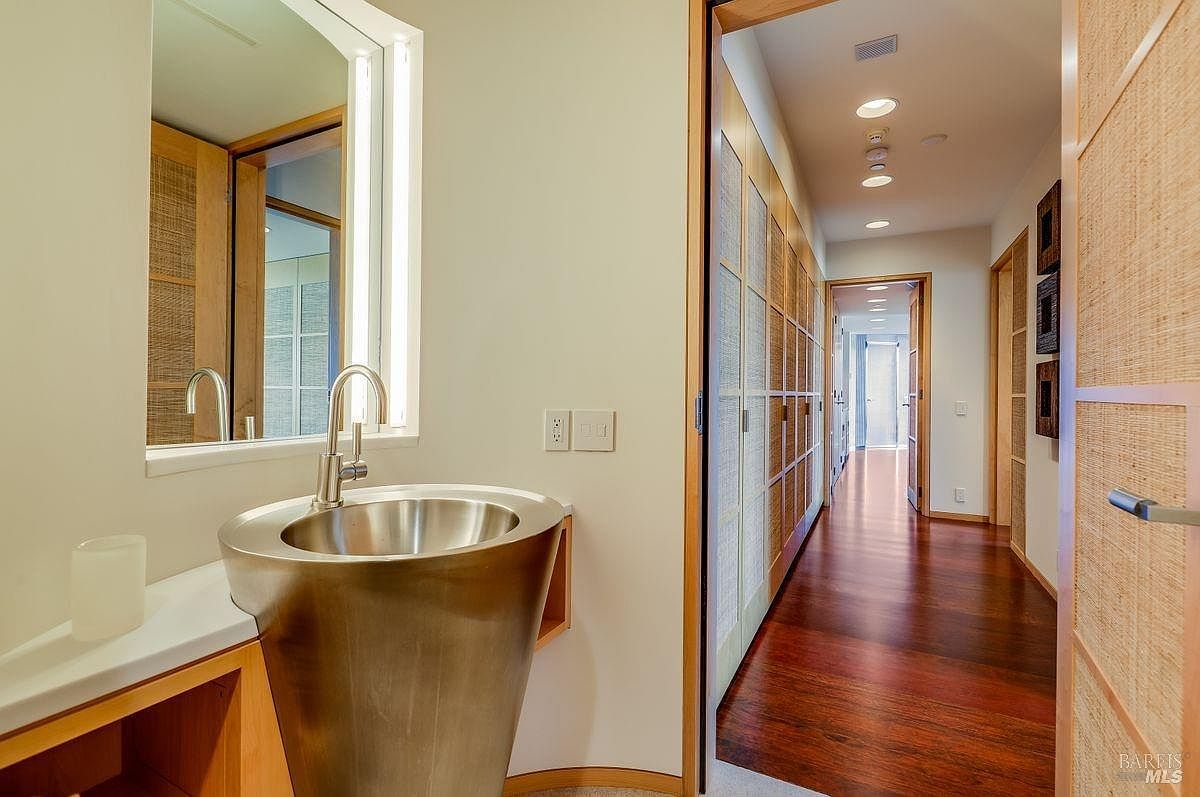 This interior view captures a modern powder room featuring a unique, conical metallic pedestal sink and a wall-mounted mirror with integrated vertical lighting. To the right, a hallway with rich wood flooring and textured wall panels leads toward a bright, open space at the end of the corridor. The design aesthetic is minimalist and sophisticated, utilizing natural textures and clean lines to create a serene atmosphere.