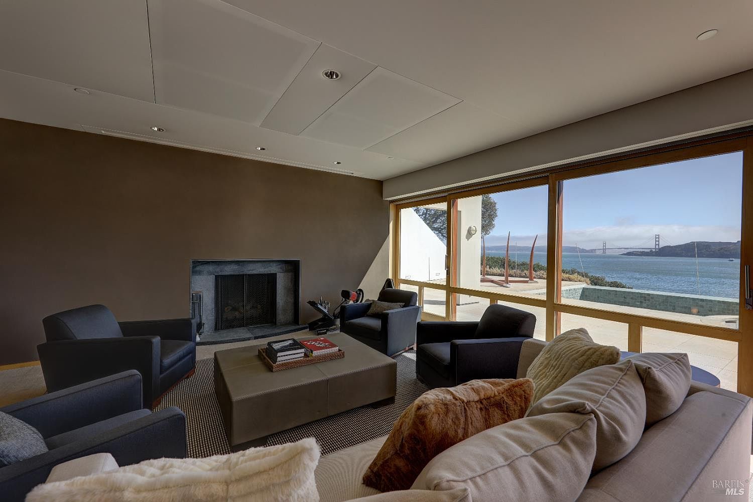 This sophisticated living room features a neutral color palette with dark armchairs arranged around a large, square ottoman. A stone-clad fireplace serves as a focal point on the solid-colored wall, while expansive windows provide a stunning, unobstructed view of the bay and the Golden Gate Bridge. The space is designed for comfort and relaxation, blending contemporary furniture with a breathtaking natural backdrop.