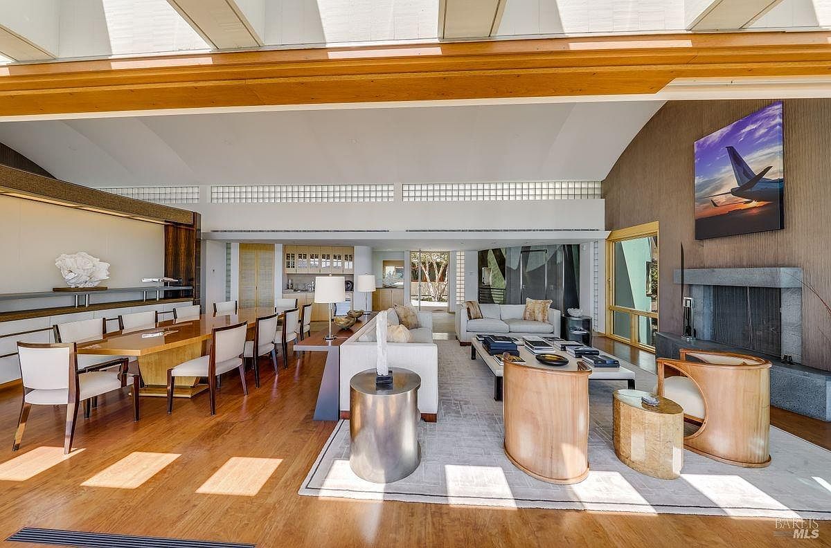 This expansive, open-concept living and dining area features warm wood flooring and a vaulted ceiling with exposed beams. The space includes a formal dining set, a comfortable seating area with a light-colored sofa, and a stone-clad chimney breast housing a fireplace. Large windows and glass doors provide a seamless connection to the outdoors, creating a bright and airy atmosphere with a sophisticated, modern aesthetic.