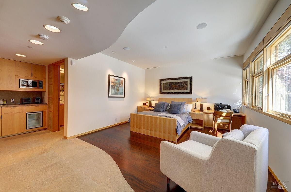 This spacious primary bedroom features a warm, contemporary design with dark wood flooring and a neutral color palette. The room includes a bed centered against a wall with a framed piece of art, a dedicated workspace with a desk and chair, and an integrated kitchenette area with light wood cabinetry. Large windows along the right wall provide ample natural light, while recessed lighting illuminates the curved ceiling architecture.