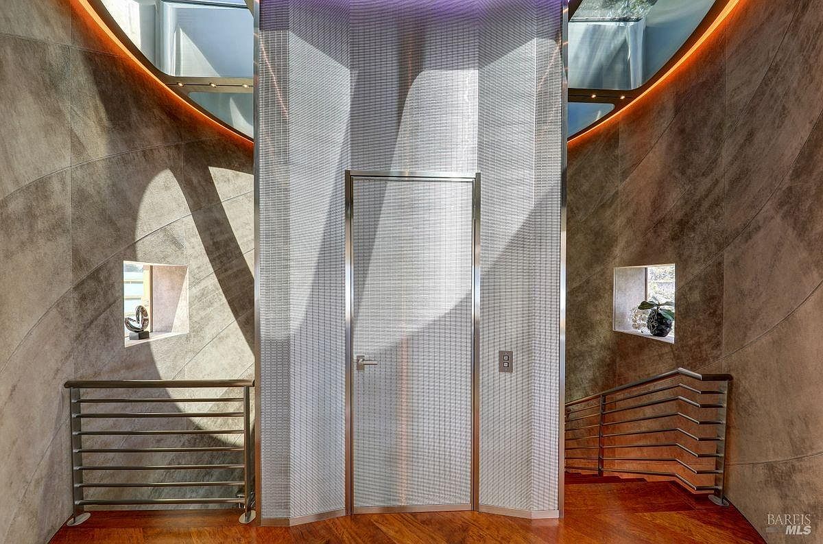 This interior space features a central elevator enclosed in a textured, metallic-finish wall, flanked by curved walls clad in large, neutral-toned stone tiles. A wooden staircase with a sleek metal railing descends to the right, while a matching railing protects the landing on the left. Small, recessed niches with decorative items add visual interest, and the curved architecture is highlighted by warm, indirect cove lighting along the ceiling line.