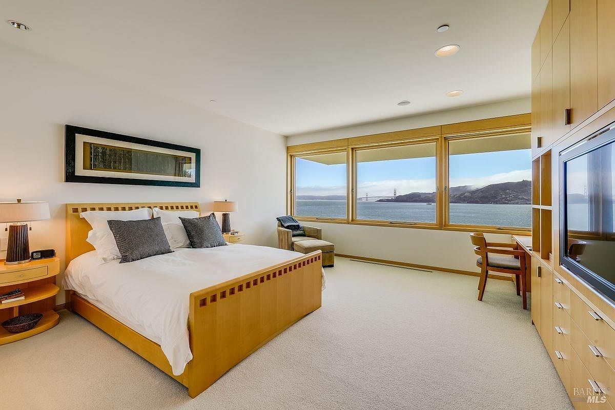 This spacious primary bedroom features a light wood bed frame and matching nightstands, complemented by a neutral color palette and plush carpeting. A wall of large windows provides an expansive, panoramic view of the bay and the Golden Gate Bridge, filling the room with natural light. To the right, a built-in light wood cabinetry unit incorporates a television and a dedicated desk area, creating a functional and cohesive living space.