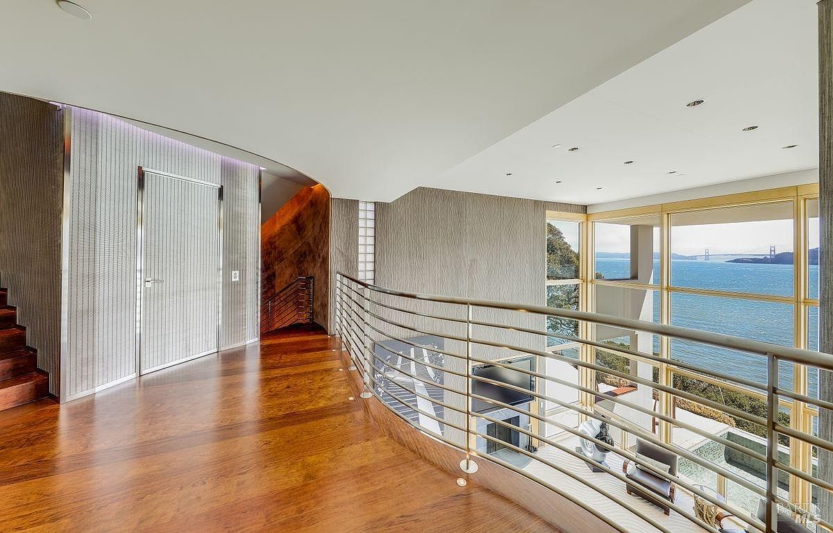 This interior view captures a curved mezzanine hallway overlooking a lower living area, characterized by rich wood flooring and a sleek metal railing. Large windows provide an expansive, unobstructed view of the bay and the Golden Gate Bridge, flooding the space with natural light. The architectural design features a curved wall with textured paneling and a modern, minimalist aesthetic.