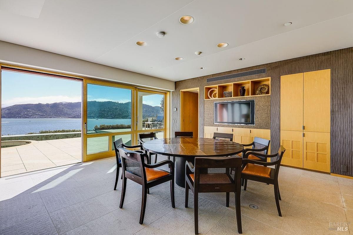This elegant dining room features a circular wooden table surrounded by dark-framed chairs with leather-toned seating. A wall-mounted television is integrated into a custom light wood cabinetry unit with open shelving, while large sliding glass doors provide a panoramic view of the water and distant hills. The room is finished with neutral stone flooring and recessed ceiling lighting, creating a sophisticated and airy atmosphere.