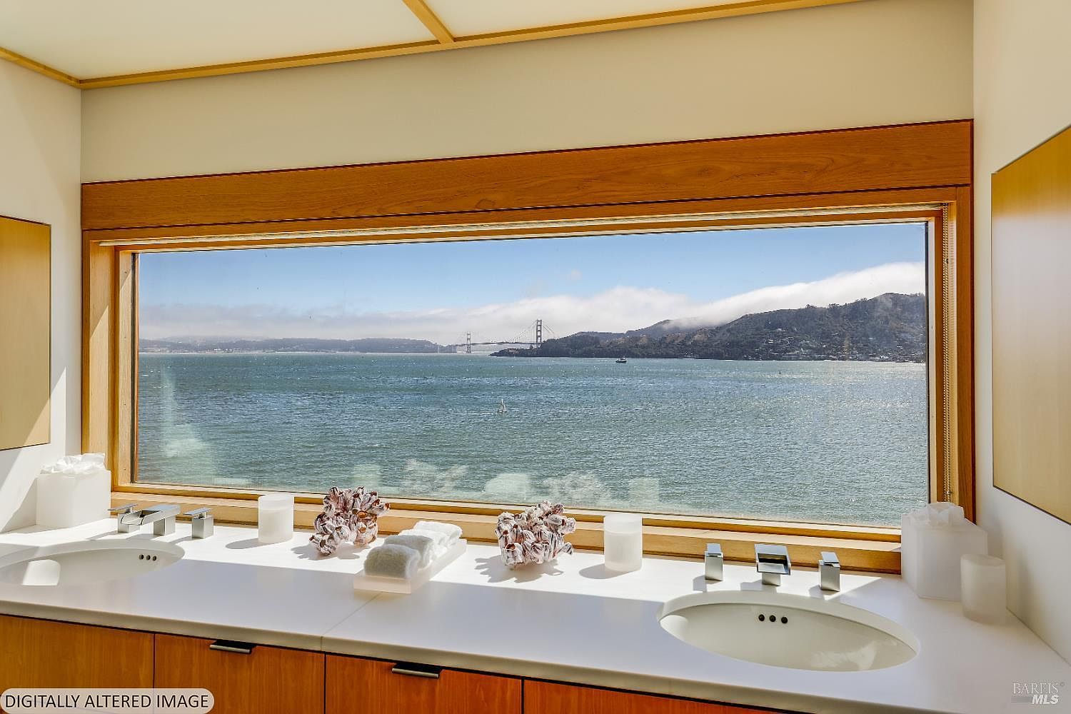 This primary bathroom features a dual-sink vanity with a clean white countertop and light wood cabinetry, positioned directly beneath a wide, horizontal window. The window provides a panoramic, unobstructed view of the bay and the Golden Gate Bridge, creating a serene and luxurious atmosphere. The space is bright and minimalist, emphasizing the connection between the interior design and the dramatic waterfront scenery.