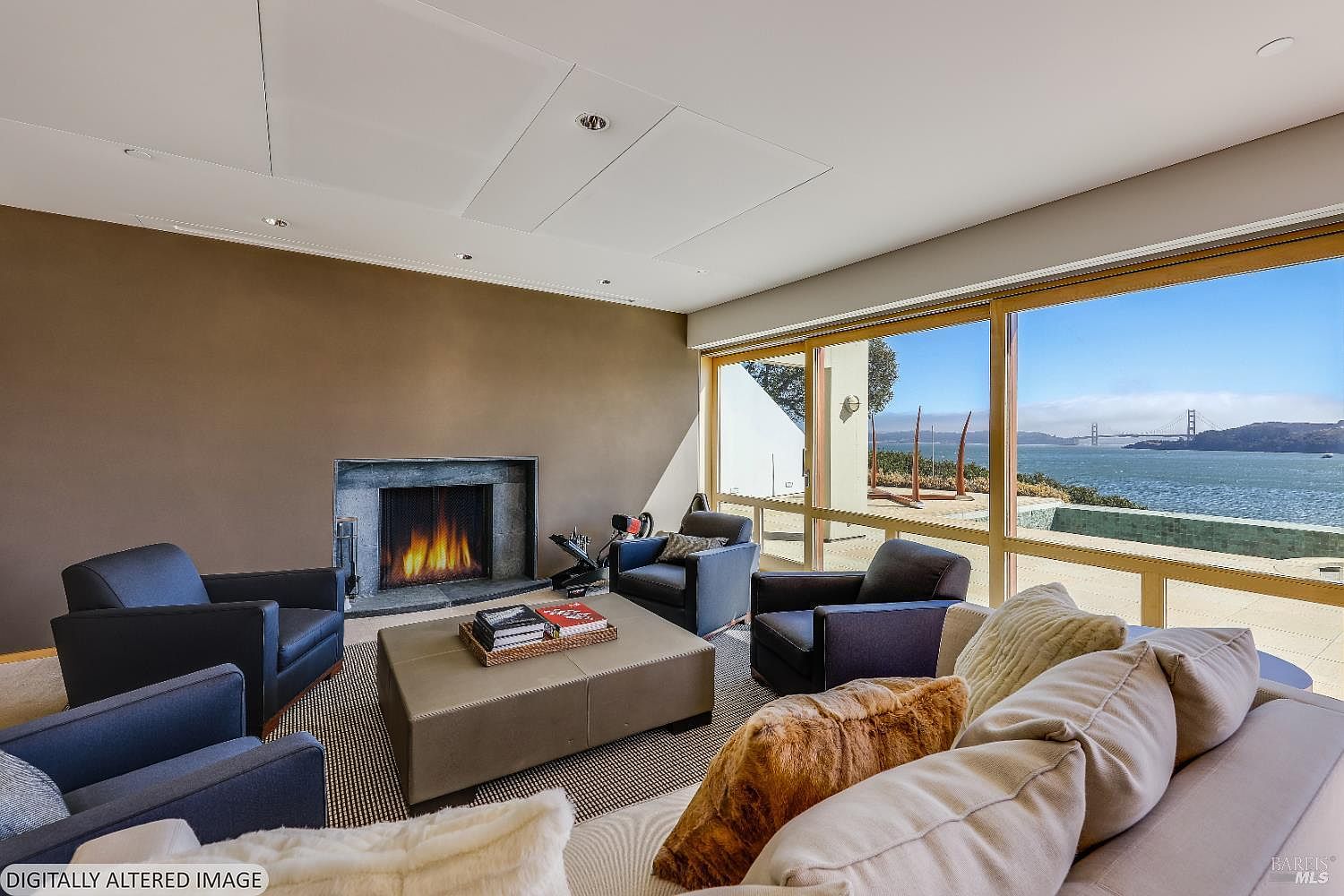 This sophisticated living room features a neutral color palette and a large, comfortable seating area centered around a square ottoman and a fireplace with a dark stone surround. Expansive windows provide a panoramic view of the bay and the Golden Gate Bridge, bathing the room in natural light. The space is designed for relaxation and entertaining, blending modern comfort with a stunning waterfront backdrop.