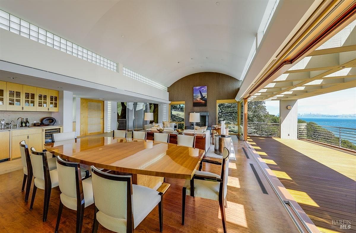 This expansive open-concept living and dining area features a vaulted ceiling and a seamless transition to a large outdoor deck through retractable glass walls. The space includes a dedicated dining area with a large wooden table, a wet bar with light wood cabinetry, and a comfortable seating area centered around a fireplace. The room offers panoramic views of the water and distant city skyline, creating a bright and airy atmosphere with a sophisticated, modern aesthetic.