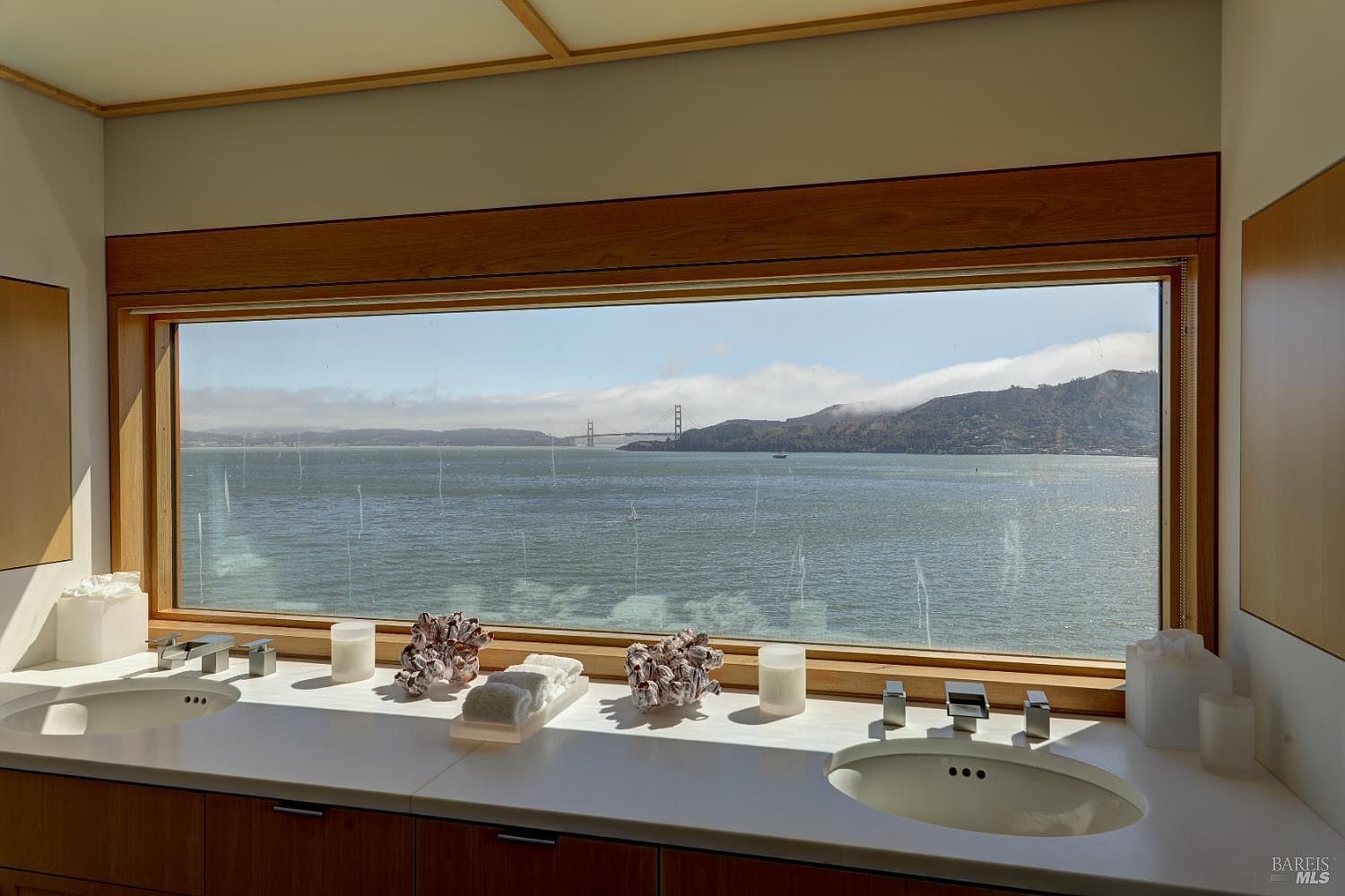 This primary bathroom features a dual-sink vanity with a clean white countertop and light wood cabinetry, positioned directly beneath a wide, panoramic window. The expansive window offers a stunning, unobstructed view of the bay and the Golden Gate Bridge, creating a serene and luxurious atmosphere. The minimalist design is accented by decorative coral pieces and simple glass candle holders, emphasizing the connection between the interior space and the scenic outdoor landscape.