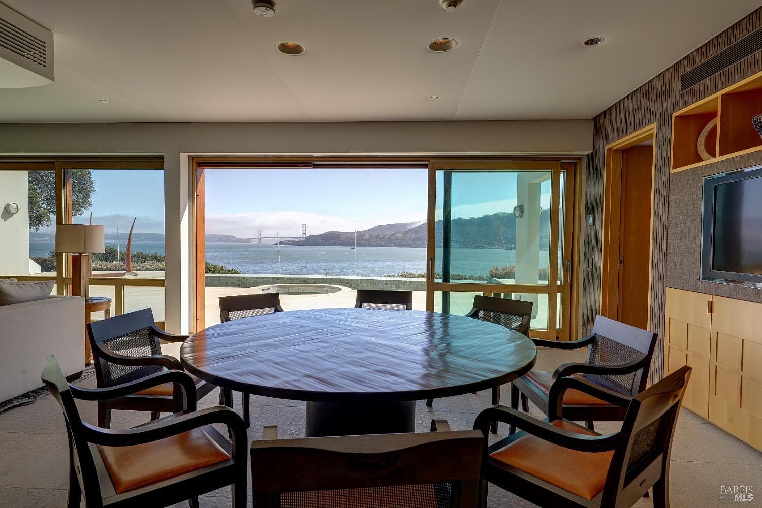 This dining room features a large, circular dark wood table surrounded by six chairs with woven backrests and tan cushions. The room is defined by expansive sliding glass doors that provide a panoramic view of the bay and the Golden Gate Bridge. The interior design is minimalist and contemporary, with recessed lighting and a built-in media cabinet along the side wall.