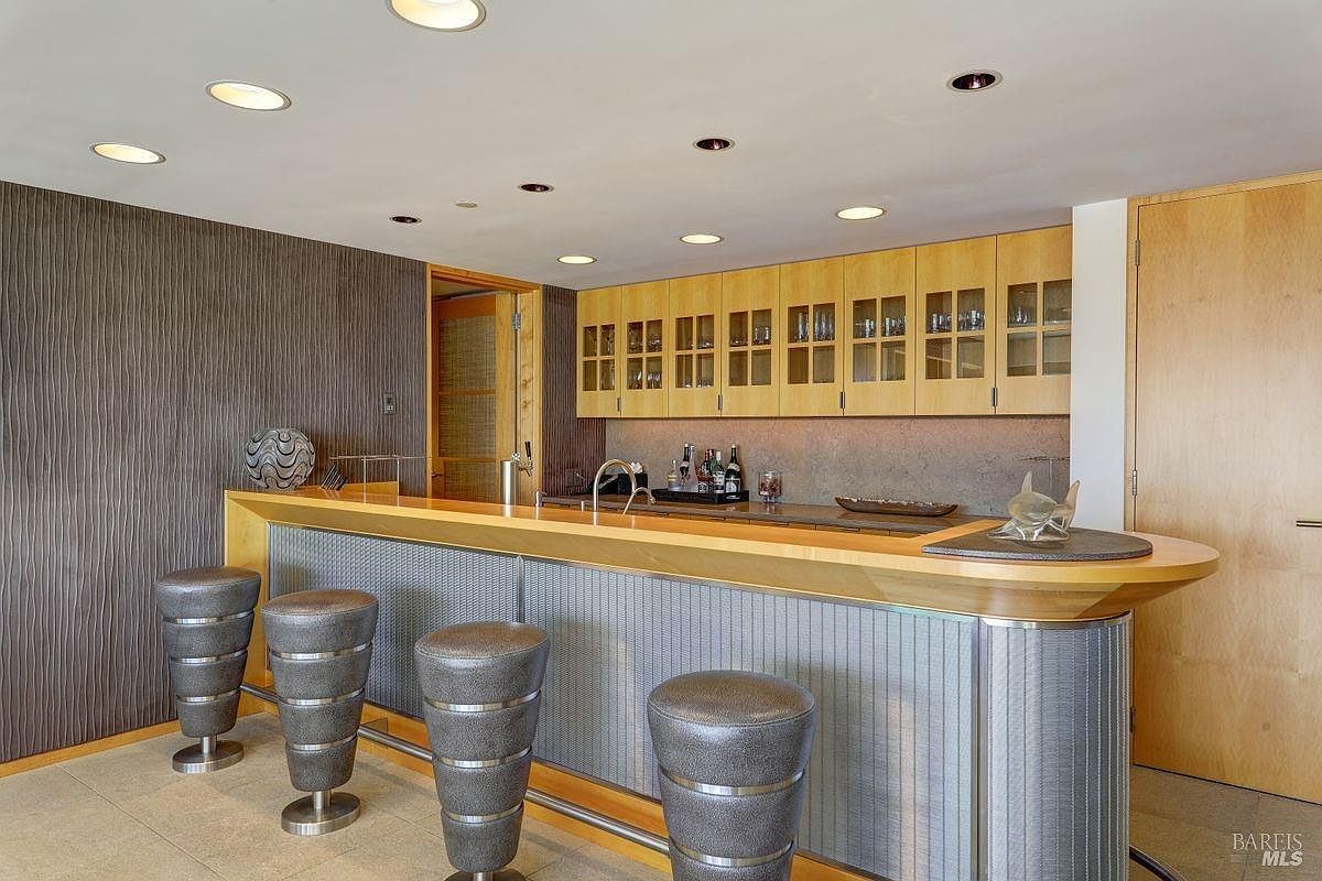 This home bar area features a long, curved counter with a light wood top and a textured, metallic-look base. Four modern, cylindrical bar stools with metallic bands are arranged along the front, while the back wall includes light wood cabinetry with glass-front doors and a neutral-toned backsplash. The space is illuminated by multiple recessed ceiling lights, creating a sleek and contemporary entertainment atmosphere.