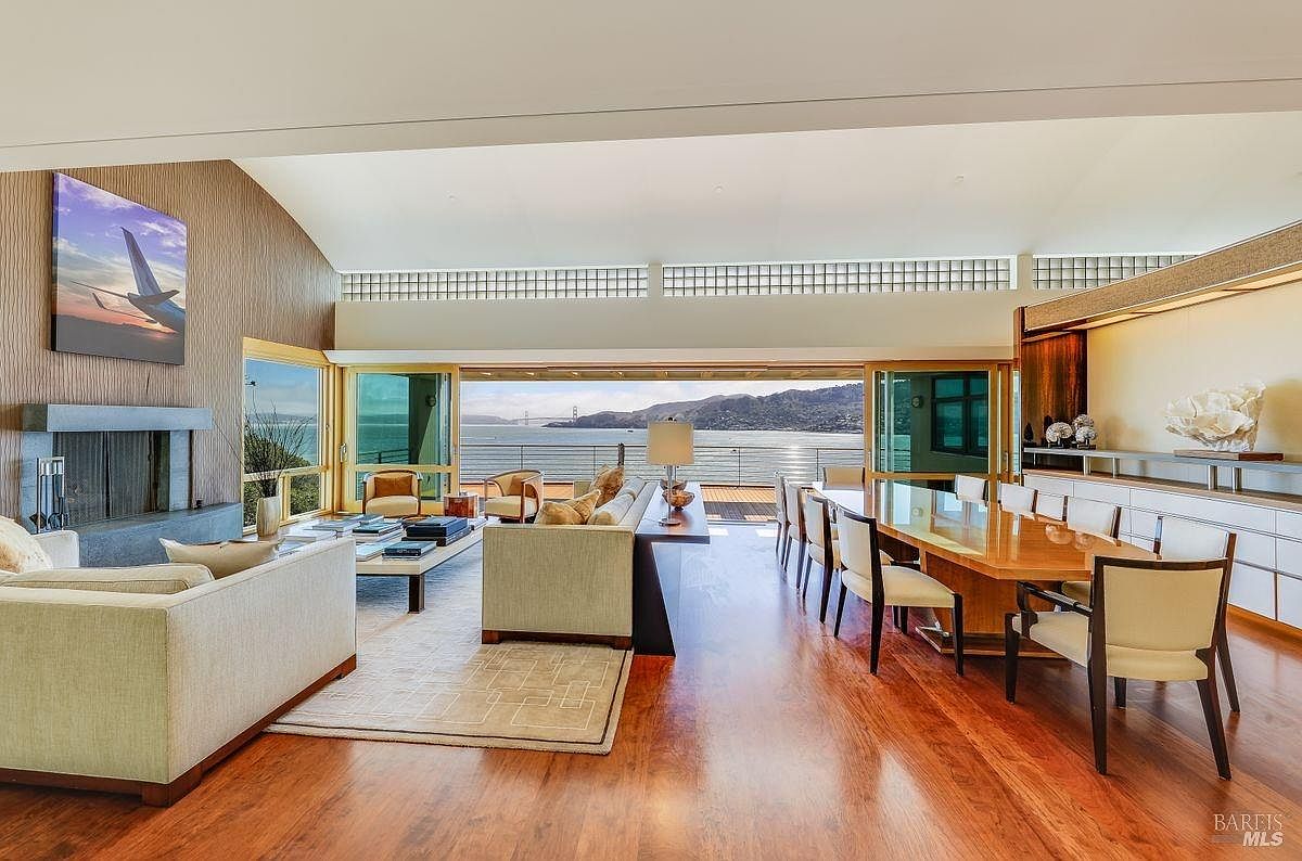 This expansive open-concept living and dining area features rich hardwood flooring and a seamless transition to an outdoor deck through large glass doors. The living space is anchored by a fireplace with a dark, minimalist surround, while the dining area is furnished with a long table and modern chairs. The room is bathed in natural light, highlighting the panoramic water views of the Golden Gate Bridge in the distance.