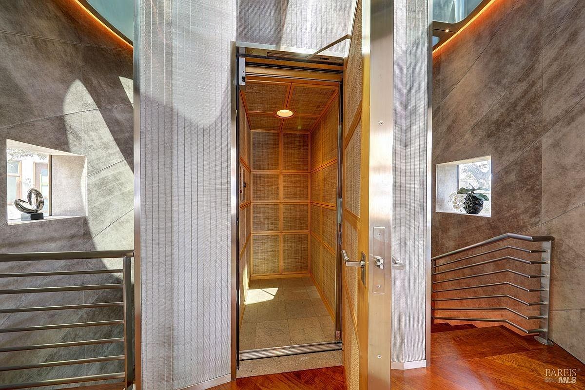 This interior view captures a modern home elevator with a textured wood-paneled interior, positioned between two staircases. The surrounding walls feature large, neutral-toned tiles and recessed lighting, while metal railings line the wooden stairs on either side. The perspective is centered on the open elevator door, highlighting the sophisticated, contemporary design of the home's circulation space.