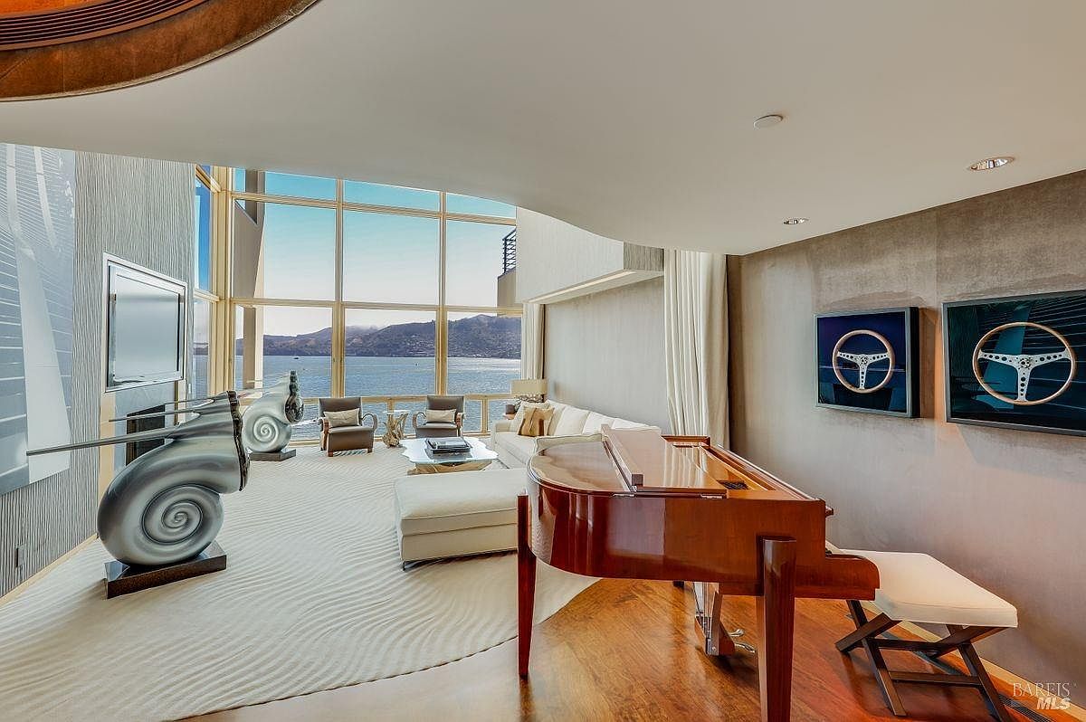 This expansive living area features a grand piano in the foreground set on rich wood flooring, transitioning into a plush, light-colored carpeted seating area. Large windows provide a panoramic view of the water and distant mountains, flooding the space with natural light. The room is decorated with unique sculptural art and framed steering wheel displays, creating a sophisticated and modern aesthetic.