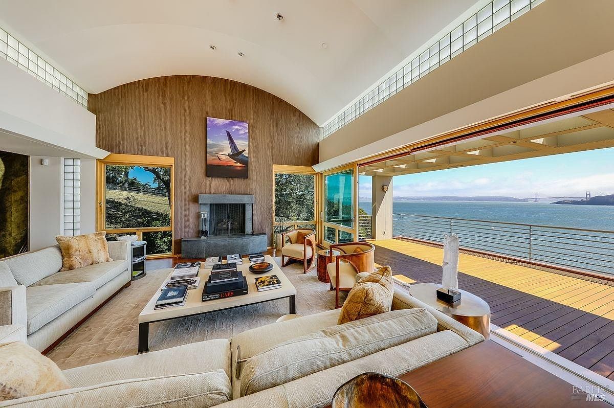 This expansive living room features a dramatic vaulted ceiling and a seamless transition to an outdoor deck through wide-opening glass doors that frame a panoramic water view. The space is anchored by a central stone fireplace with a tall surround and is furnished with comfortable, neutral-toned seating arranged around a large coffee table. The room's design emphasizes an open, airy atmosphere, blending sophisticated indoor comfort with the natural beauty of the coastal landscape.