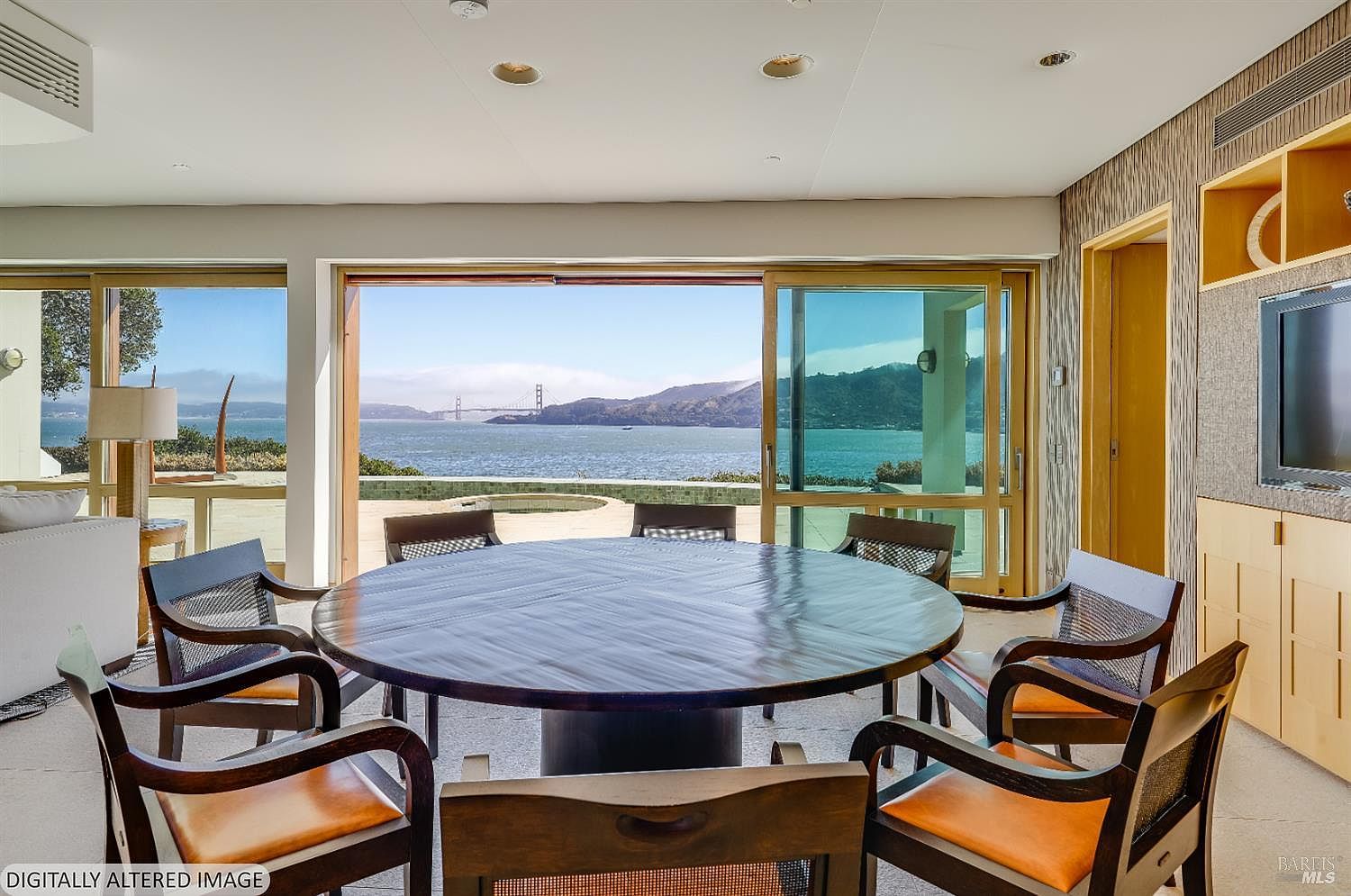 This dining room features a large, round dark wood table surrounded by six chairs with leather seats and woven backrests. The room opens up through expansive sliding glass doors to a stunning, unobstructed view of the bay and the Golden Gate Bridge. The interior aesthetic is minimalist and sophisticated, emphasizing the connection between the indoor dining space and the scenic outdoor landscape.