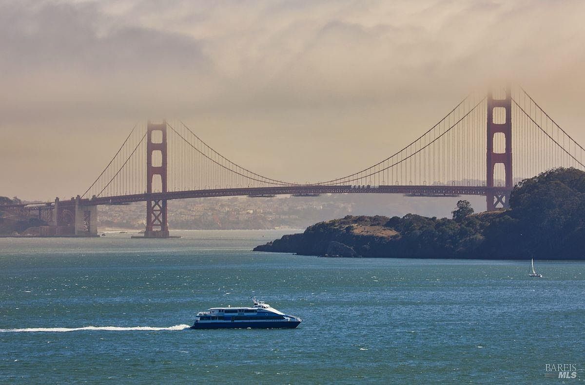 This scenic landscape shot captures the iconic Golden Gate Bridge partially shrouded in a soft, golden-hued fog. In the foreground, a passenger ferry cuts through the deep blue waters of the bay, while a small sailboat is visible near the rugged, tree-lined coastline. The perspective is a wide, elevated view that emphasizes the scale of the bridge and the tranquil maritime atmosphere of the San Francisco Bay.