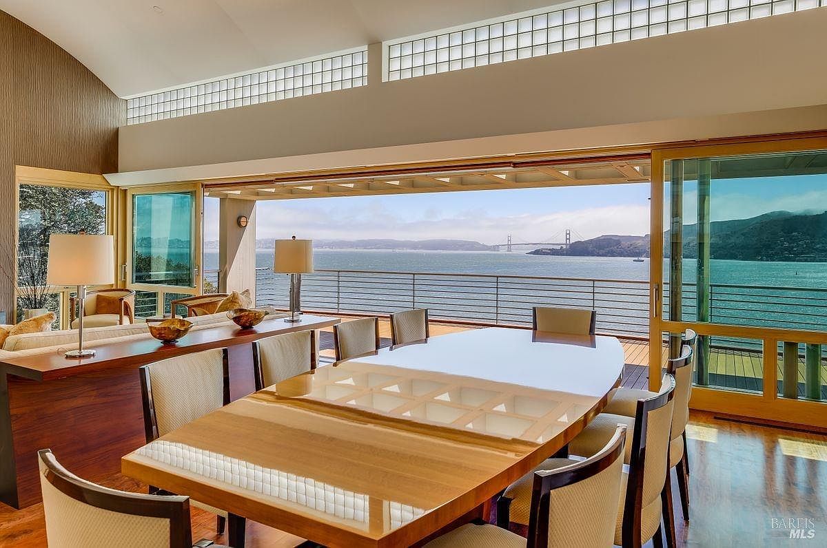This elegant dining room features a large, polished wood table surrounded by upholstered chairs, positioned to take full advantage of the panoramic bay views. The space is defined by expansive glass walls that slide open to a deck, seamlessly blending the indoor living area with the outdoor scenery, including a view of the Golden Gate Bridge. The room is accented by warm wood tones, a long console table with matching lamps, and a high, vaulted ceiling with clerestory windows that flood the area with natural light.
