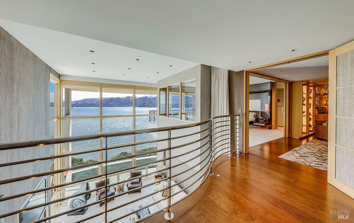 This elevated hallway features a curved metal railing that overlooks a double-height living area with expansive windows showcasing a panoramic water view. The space is finished with warm wood flooring and leads into a secondary room through a wide doorway, creating an open and airy architectural flow. The perspective is captured from the upper landing, emphasizing the dramatic verticality and the seamless connection between the interior living spaces and the outdoor scenery.