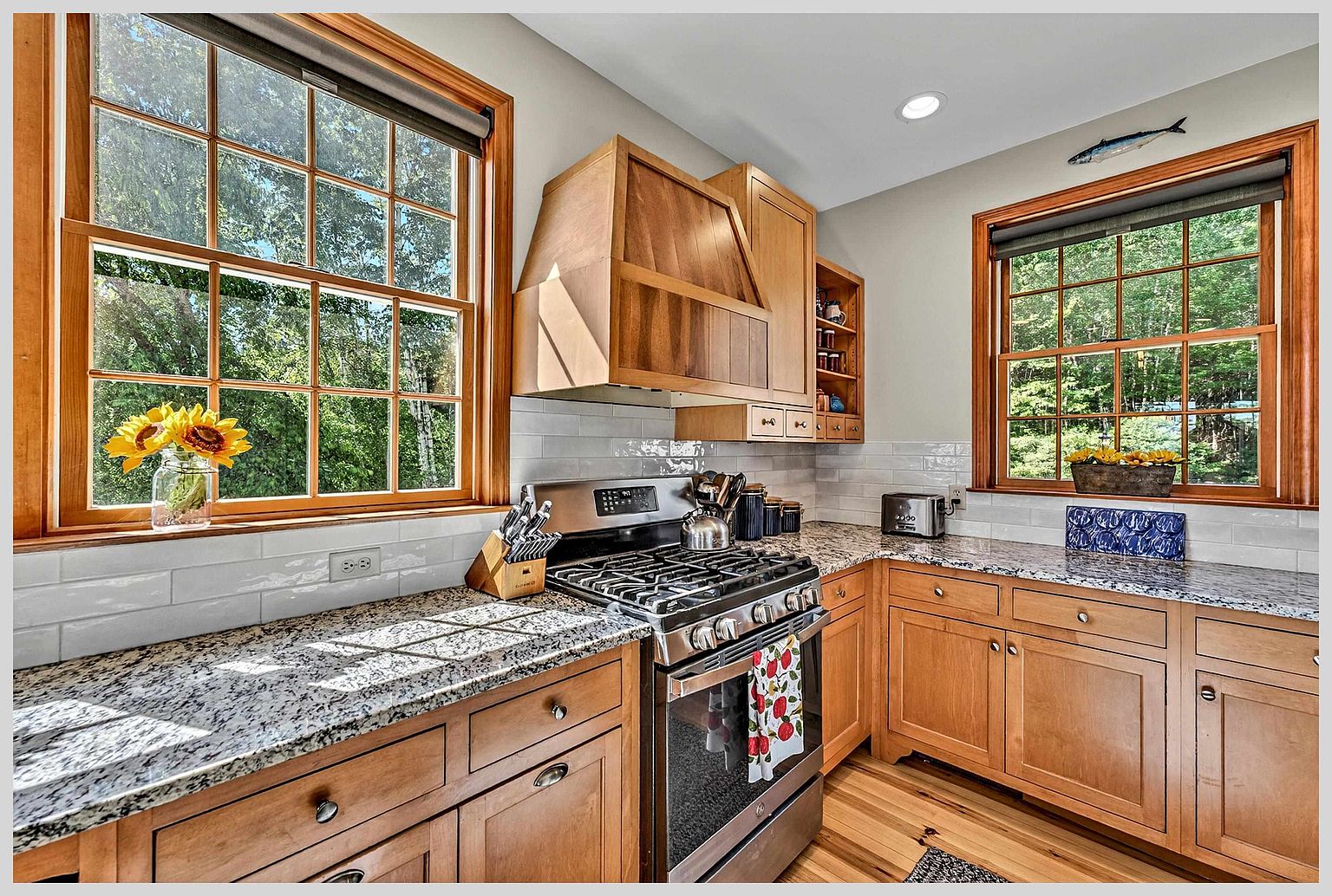 This warm and inviting kitchen features light wood cabinetry paired with speckled stone countertops and a classic white subway tile backsplash. The space is anchored by a stainless steel gas range and a custom wood-paneled range hood, while two large windows on adjacent walls flood the area with natural light. A decorative fish wall hanging and a vase of sunflowers add a touch of personality to this cozy, functional culinary space.