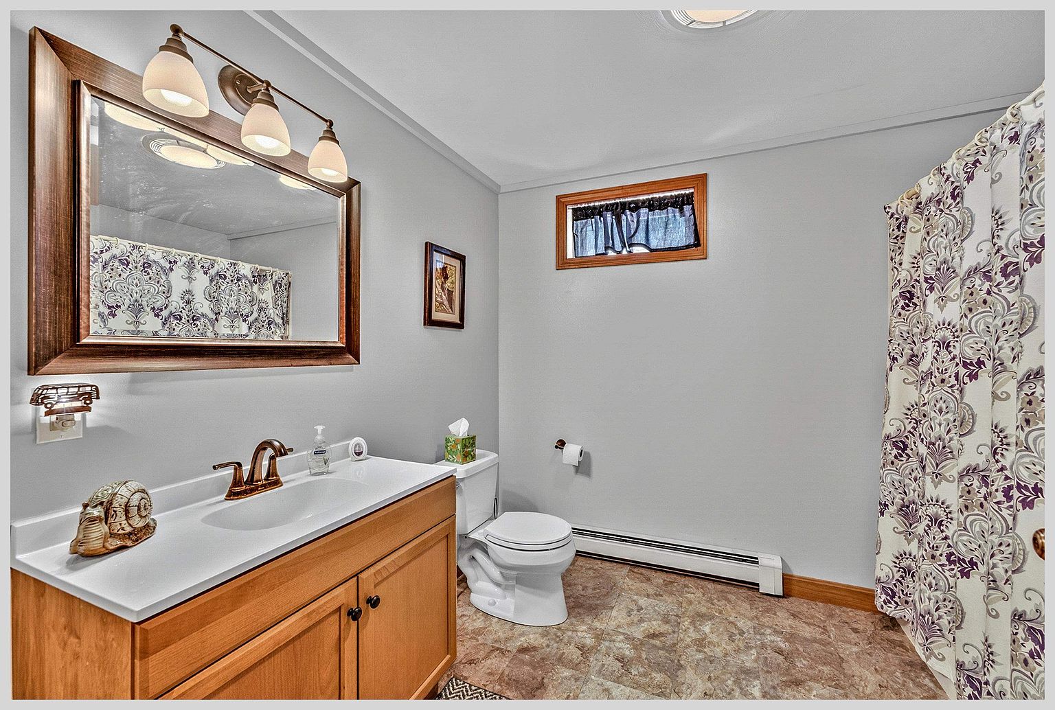 This functional bathroom features a light wood vanity with a white countertop, a large framed mirror, and a classic toilet. The space is finished with light gray walls, patterned flooring, and a shower area enclosed by a decorative floral-patterned curtain. A small window with a valance provides natural light, creating a clean and straightforward aesthetic.