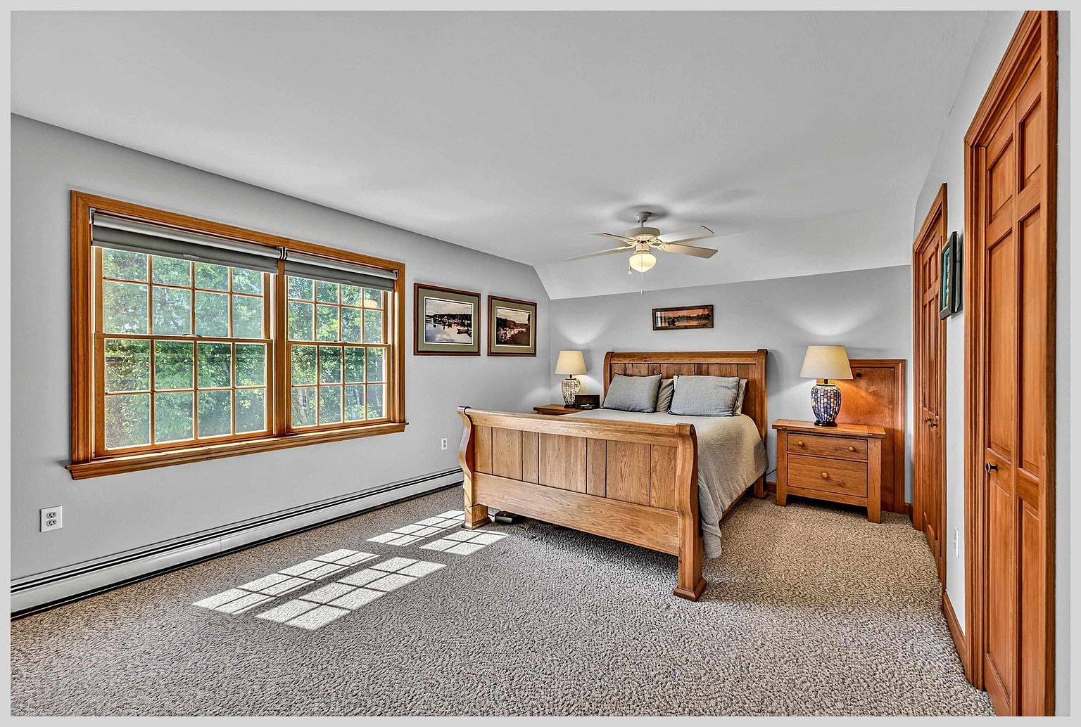 This primary bedroom features a warm, light wood bed frame as the centerpiece, flanked by matching nightstands and table lamps. Large windows on the left wall allow natural light to stream in, casting shadows across the neutral-toned carpet. The room is finished with light gray walls, a ceiling fan, and wooden doors leading to a closet, creating a cozy and traditional atmosphere.