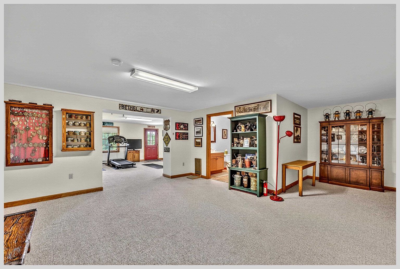 This spacious, carpeted basement area features a versatile layout with a dedicated home gym space visible through an open doorway. The room is decorated with various wall-mounted collectibles and includes a large wooden hutch, a green display shelf, and a red floor lamp. The neutral color palette and functional design suggest a comfortable, multi-purpose living area.