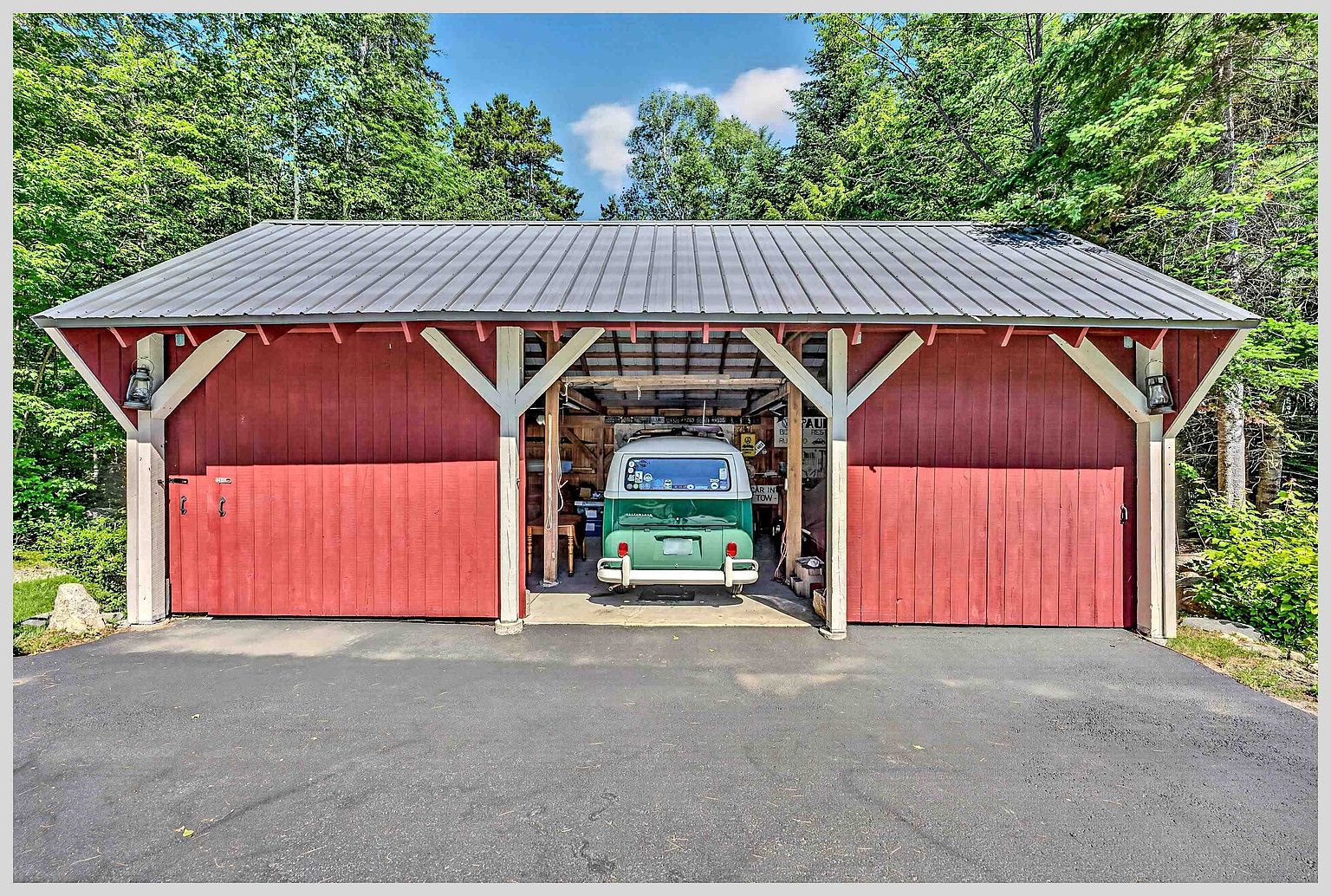 This image features a rustic, red-painted wooden garage structure with a metal roof, set against a lush, green forest backdrop. The open-bay design reveals a vintage green Volkswagen bus parked inside, adding a charming, nostalgic touch to the property. The structure is framed by sturdy wooden posts and decorative braces, creating a welcoming and well-maintained outdoor storage space.