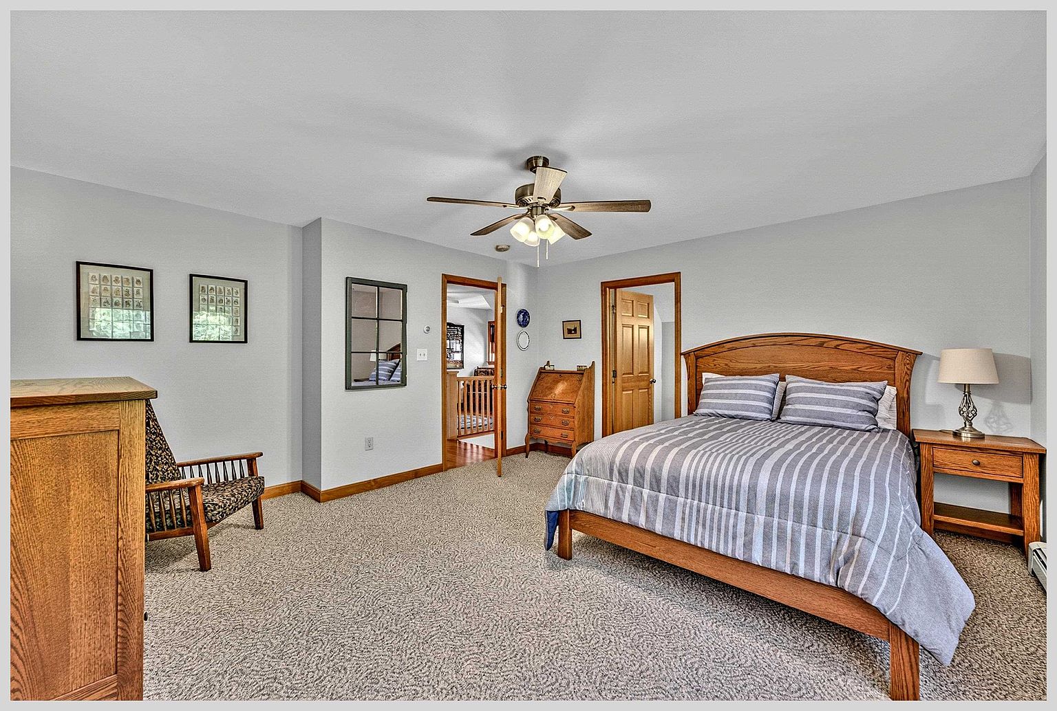 This spacious primary bedroom features a warm wood bed frame with a striped comforter and a matching nightstand with a lamp. The room is carpeted throughout and includes a wooden chair in the corner, a small secretary desk, and two framed art pieces on the wall. A ceiling fan provides overhead lighting, and the layout includes two doorways leading to other parts of the home.