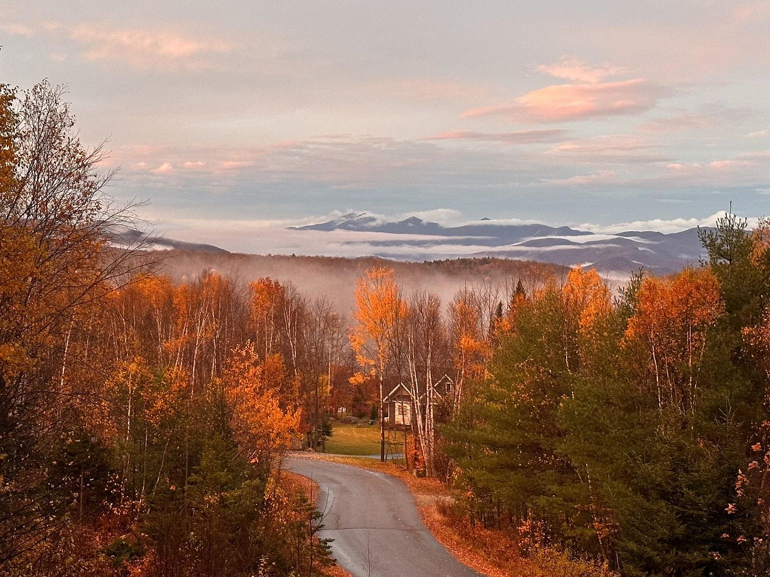 This elevated, scenic view captures a secluded property nestled within a vibrant autumn forest. A winding paved road leads toward a small, elevated cabin structure surrounded by dense, colorful foliage and a backdrop of misty, layered mountain ranges under a soft, pastel-hued sky. The perspective emphasizes the tranquil, remote nature of the setting, highlighting the integration of the home into the natural landscape.