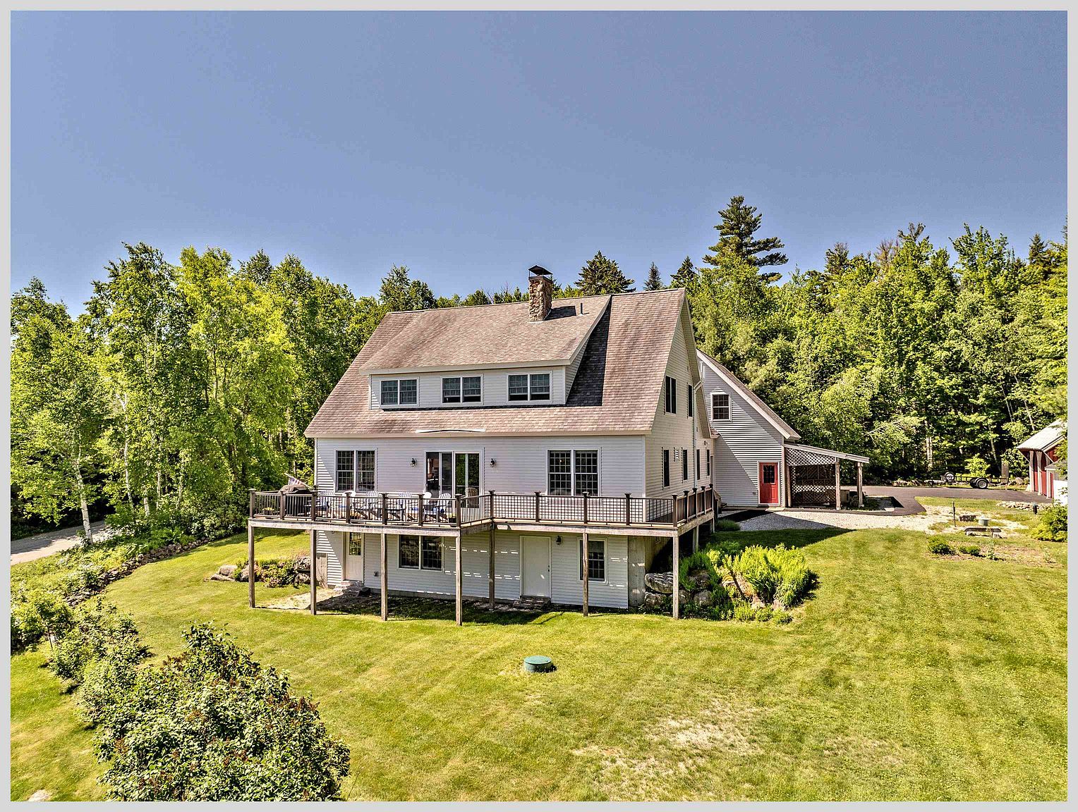 This elevated perspective captures the rear of a multi-story home featuring white siding and a prominent, expansive wooden deck that spans the width of the main level. The house includes a dormer with multiple windows, a stone chimney, and a lower-level walk-out basement area. Surrounded by a lush, mature forest, the property offers a spacious, well-maintained lawn that slopes gently away from the structure.