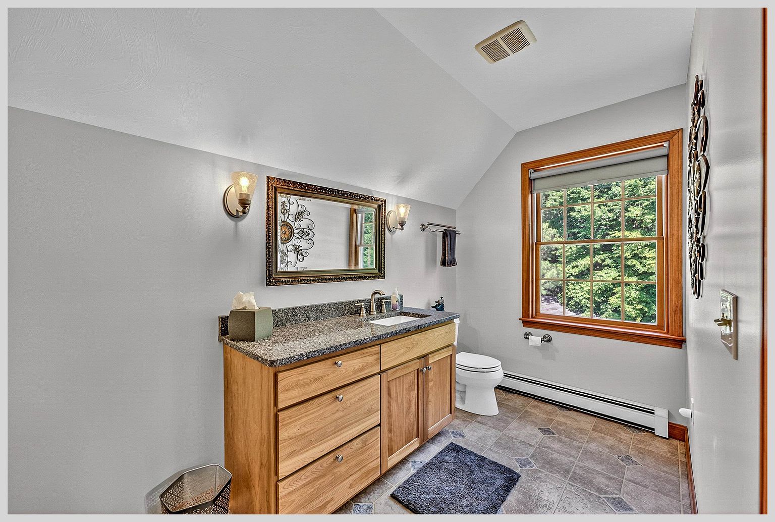 This bathroom features a light wood vanity topped with a speckled stone counter and a single undermount sink. A framed mirror hangs above the vanity, flanked by two wall-mounted light fixtures, while a window with wooden trim provides natural light. The room is finished with gray walls and patterned tile flooring, creating a functional and neutral space.