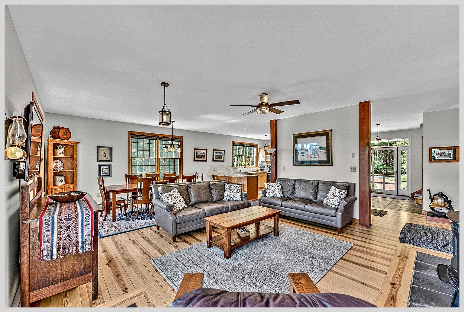 This open-concept living and dining area features warm light wood flooring and a neutral color palette, creating a welcoming and spacious atmosphere. The room is furnished with two dark leather sofas, a wooden coffee table, and a dining set, all arranged to maximize the flow between the living space and the adjacent kitchen. Large windows allow natural light to fill the room, while decorative accents like a patterned runner and framed artwork add character to the home.