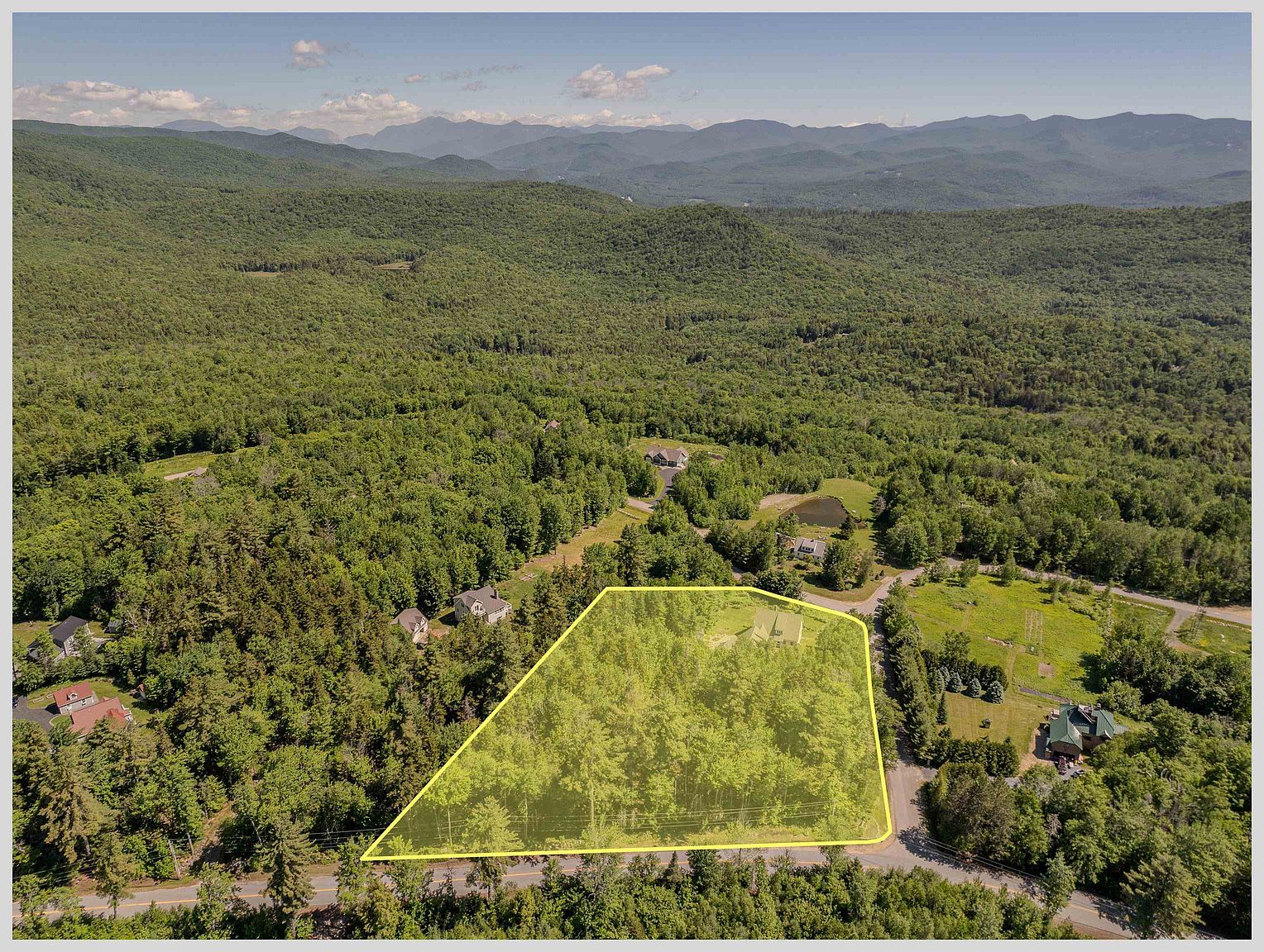 This high-angle aerial view showcases a wooded residential lot highlighted with a yellow boundary overlay, situated in a lush, mountainous landscape. The property features a dense forest canopy, a small clearing with a structure, and direct access to a paved road, all set against a backdrop of rolling hills and distant mountain ranges. The perspective provides a clear sense of the property's size, topography, and its serene, secluded location within a natural environment.