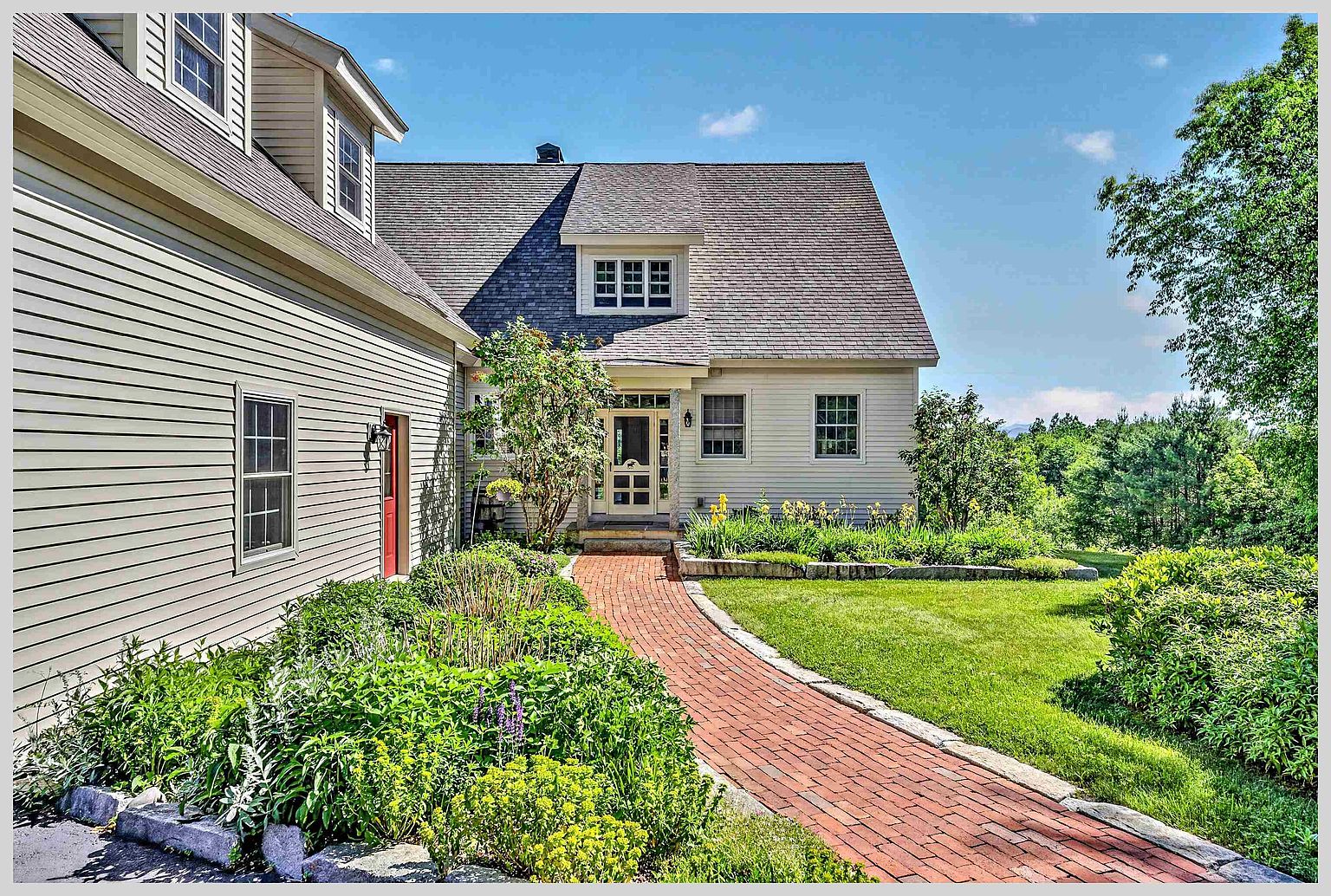 This inviting home features a classic, light-colored siding exterior with a charming brick walkway leading to the front entrance. The scene is framed by lush green landscaping, including a well-manicured lawn and vibrant garden beds, under a clear blue sky. The perspective captures the welcoming approach to the residence, highlighting its traditional architectural style and peaceful, scenic setting.