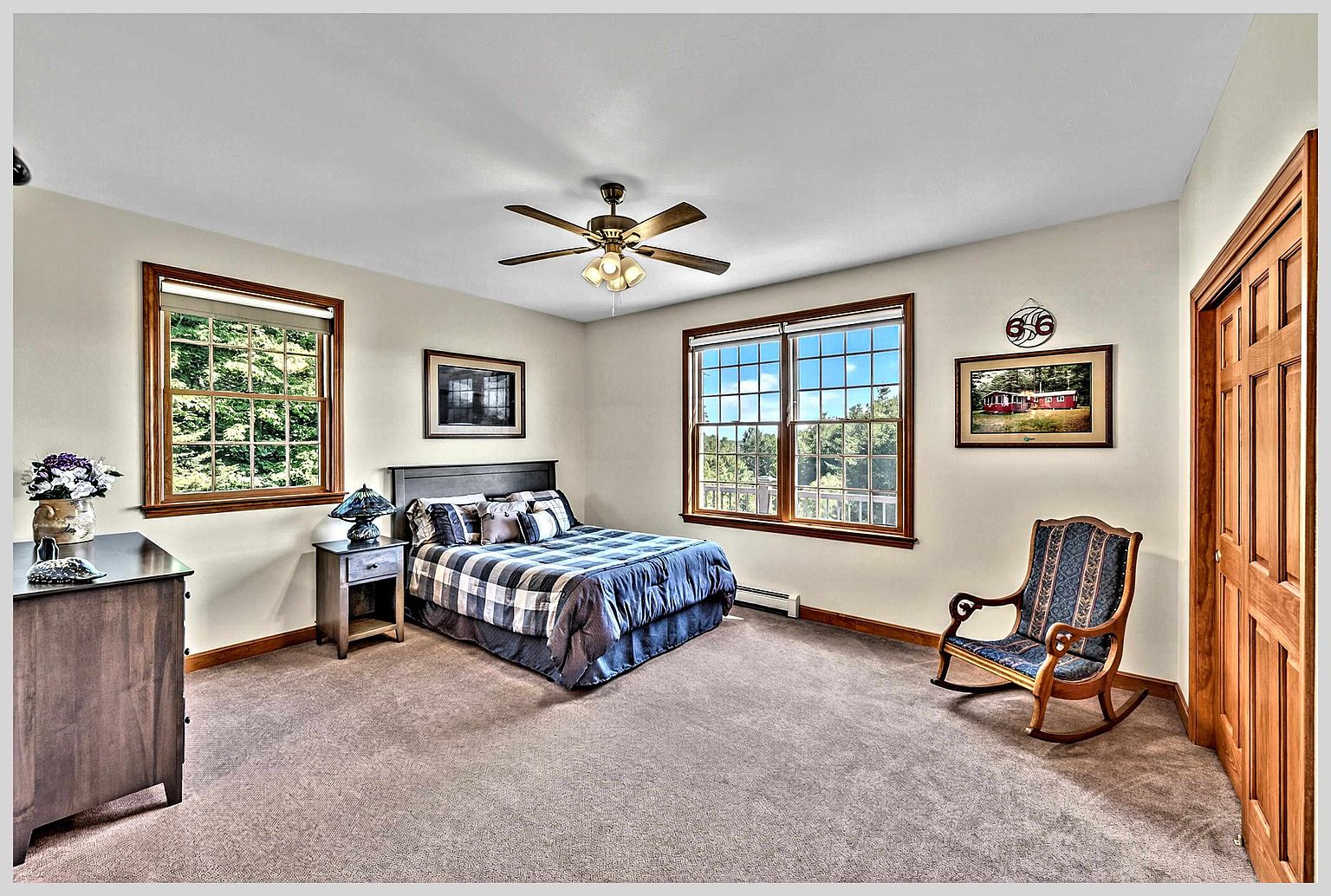 This spacious bedroom features neutral walls, plush carpeting, and a ceiling fan with integrated lighting. The room is furnished with a bed dressed in a blue and white plaid comforter, a wooden nightstand, and a classic rocking chair positioned near a wooden door. Large windows on two walls provide ample natural light and views of the surrounding trees, creating a bright and comfortable atmosphere.