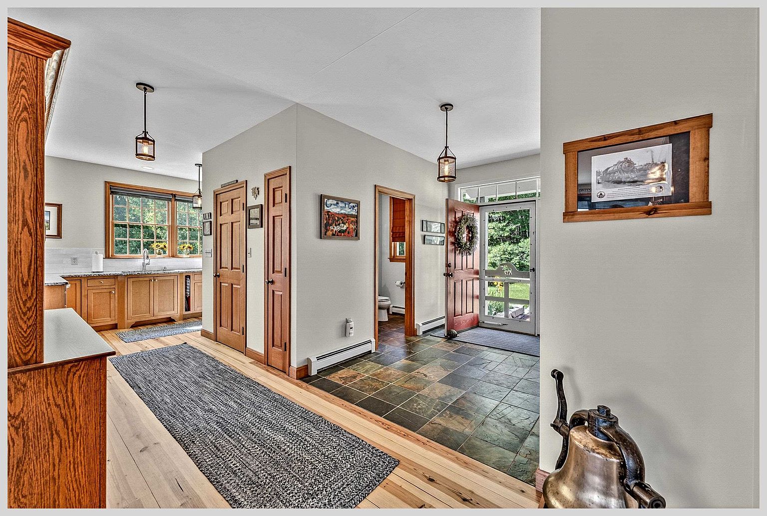This interior view captures a welcoming entryway and hallway that transitions into a kitchen area. The space features a combination of light wood flooring and dark, multi-toned slate tiles, with warm-toned wooden cabinetry and doors throughout. A decorative brass bell sits in the foreground, while a framed train print hangs on the wall, creating a cozy and inviting atmosphere.