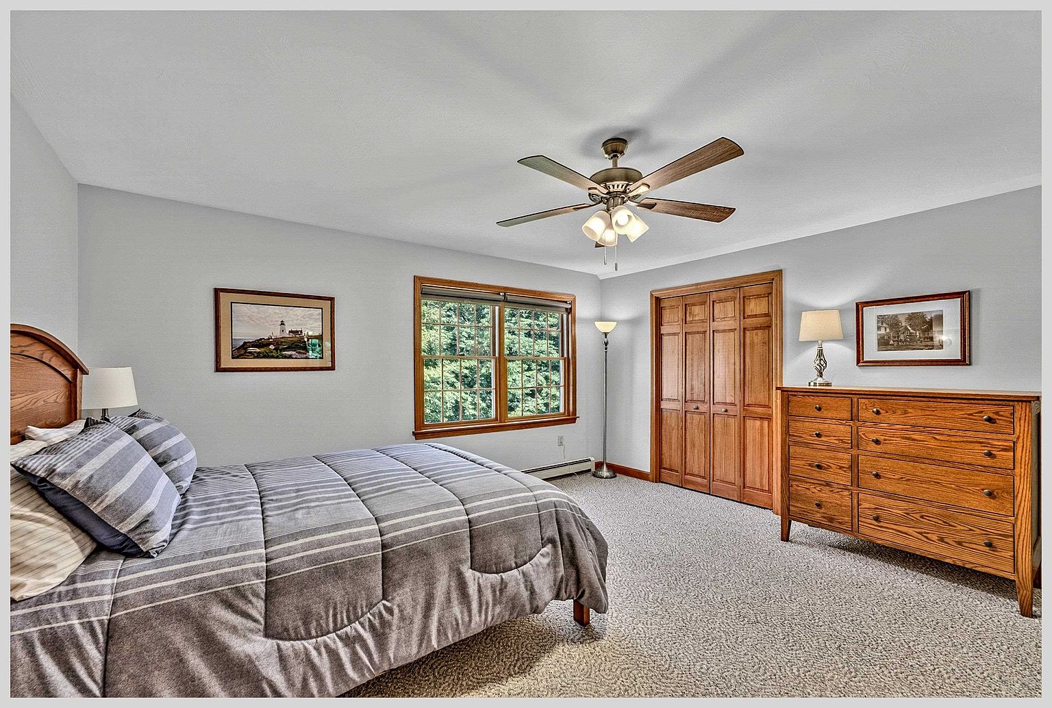 This bedroom features light-colored walls, neutral carpeting, and a wooden bed frame dressed in a striped comforter. A wooden dresser with a matching lamp sits against the wall, while a ceiling fan with integrated lighting provides overhead illumination. A window with wooden trim offers a view of the surrounding trees, creating a calm and traditional atmosphere.